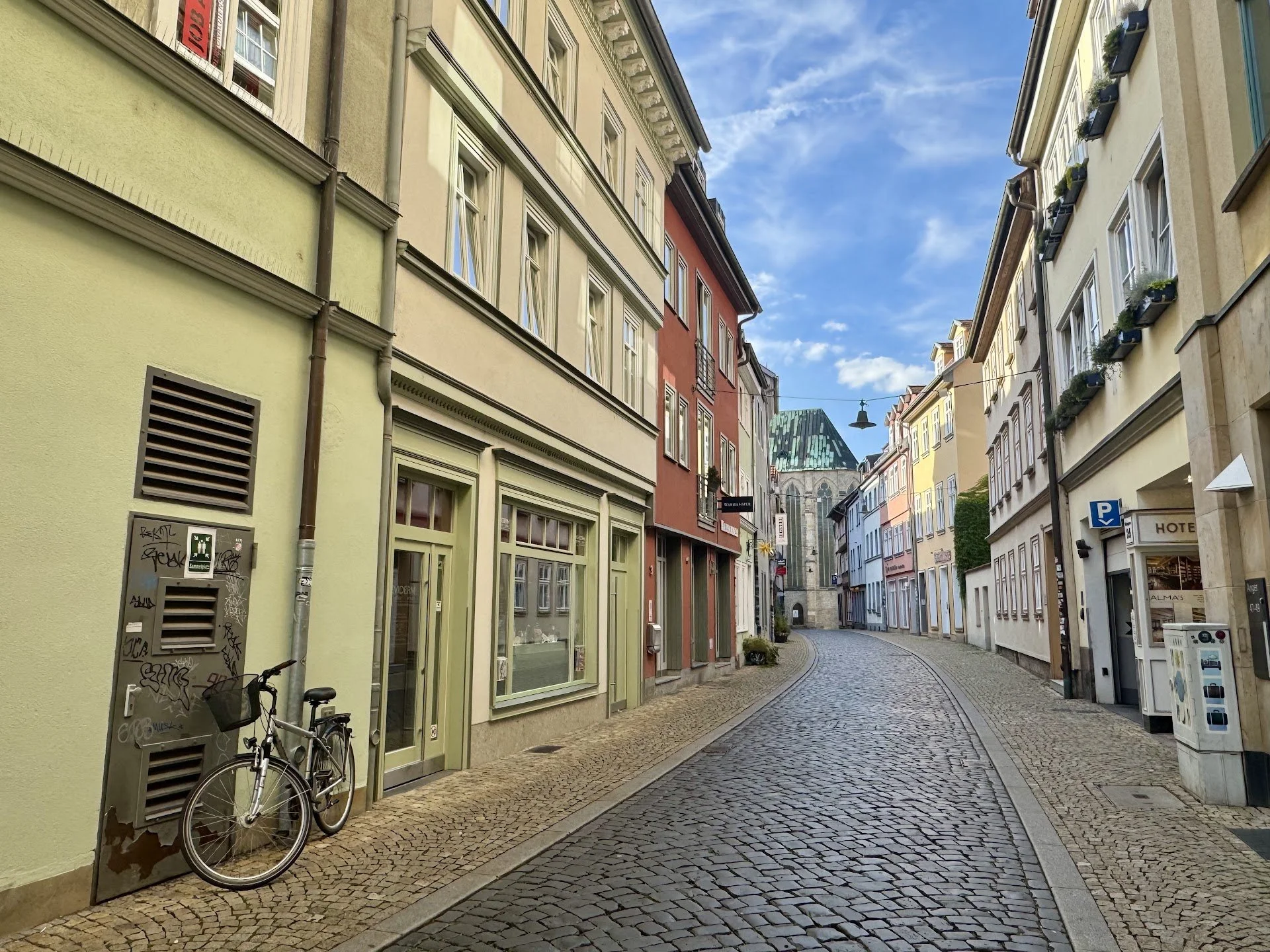 Colorful European street with cobblestone sidewalk, pastel-colored buildings, bicycles, and a church with a green copper roof in the background under a partly cloudy blue sky.