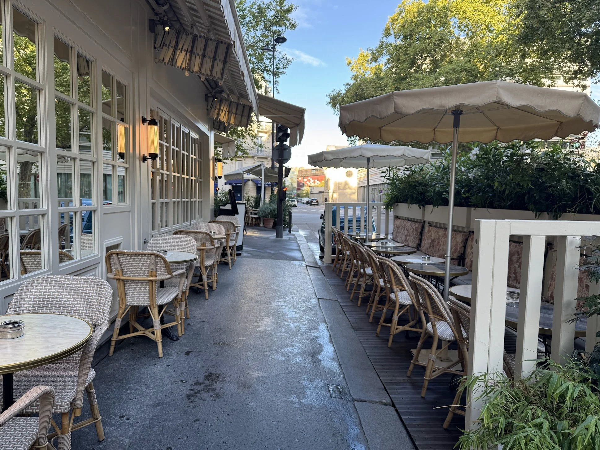 Empty outdoor restaurant patio with wicker chairs and tables, large umbrellas, and green plants, with a city street in the background.