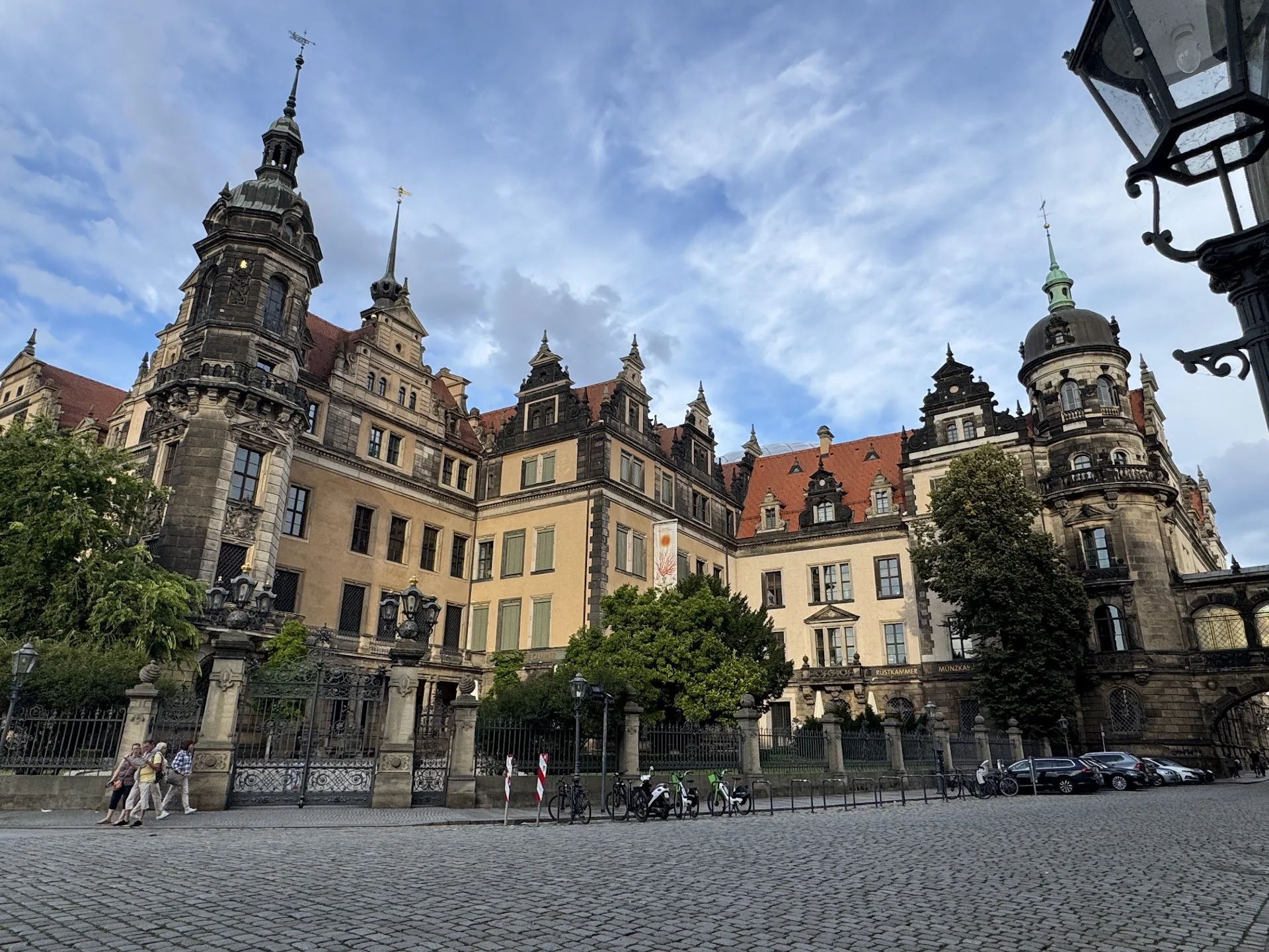 Historic European-style building with multiple towers and ornate architecture, trees in the foreground, and a cobblestone street with parked bicycles and pedestrians.