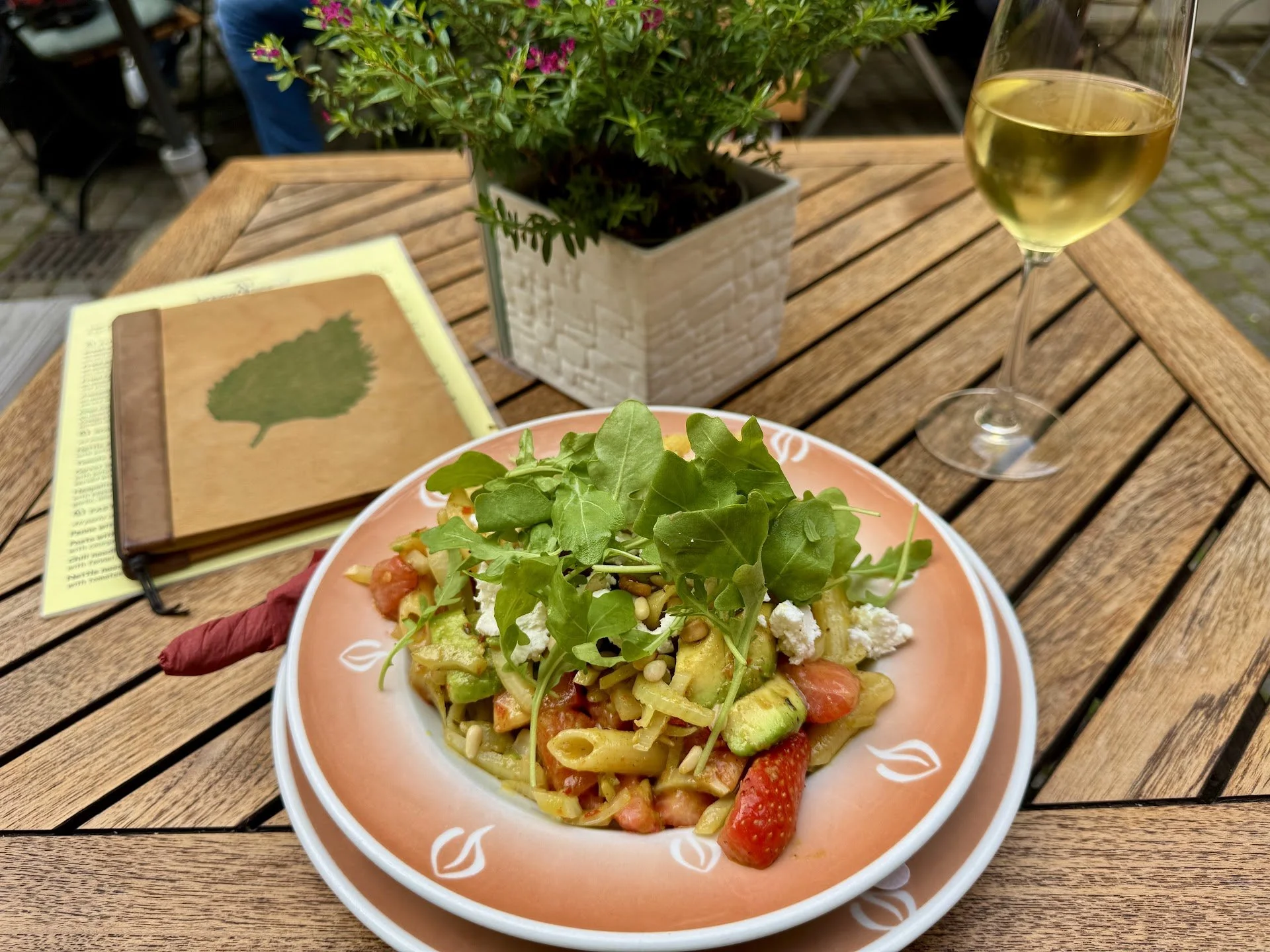 A plate of pasta topped with arugula, cherry tomatoes, zucchini, and feta cheese on an outdoor wooden table. A glass of white wine is nearby, and there is a potted plant and a menu on the table.