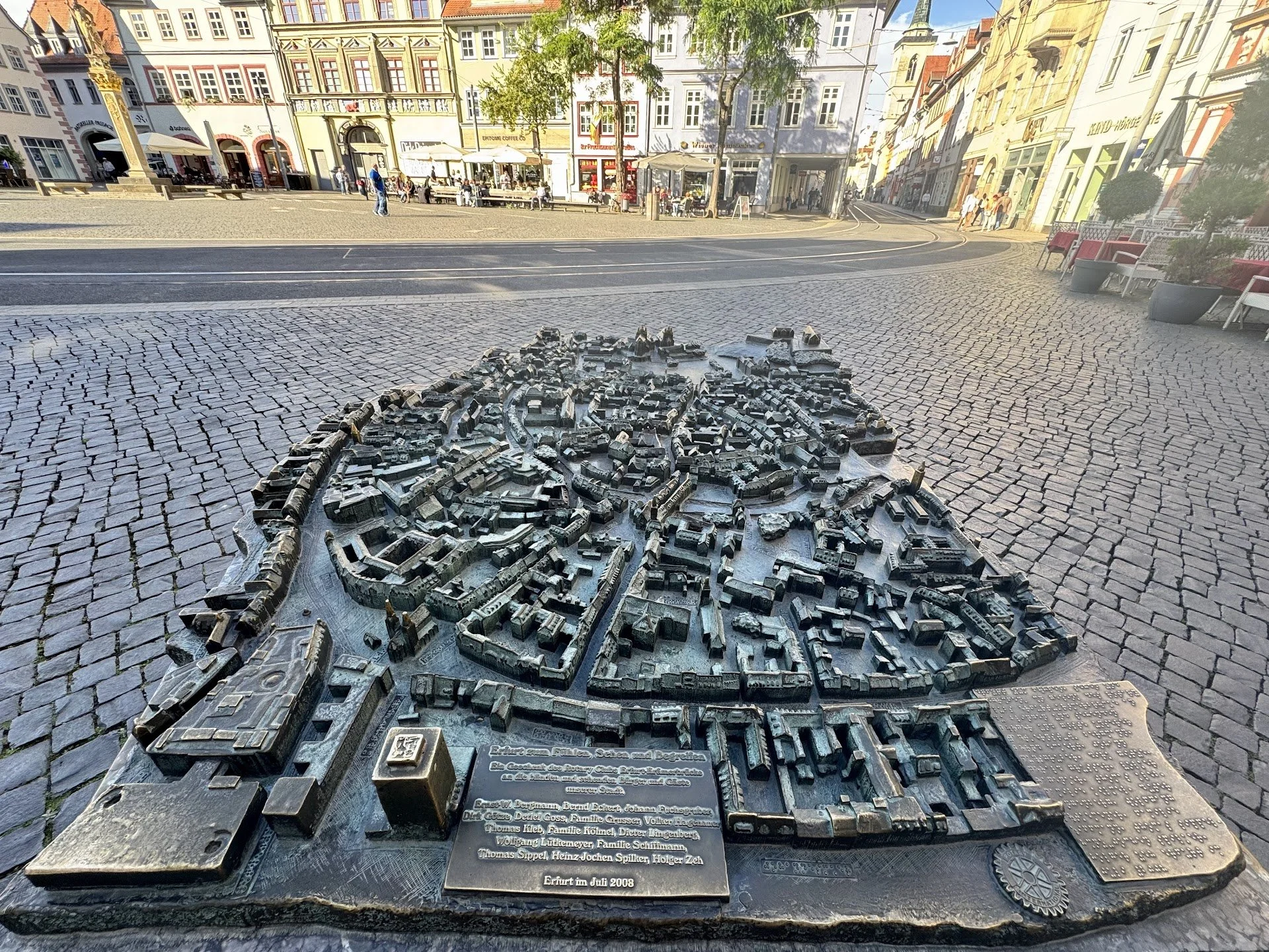A detailed map sculpture of the city in front of colorful historic buildings on a cobblestone square, with benches and people walking.