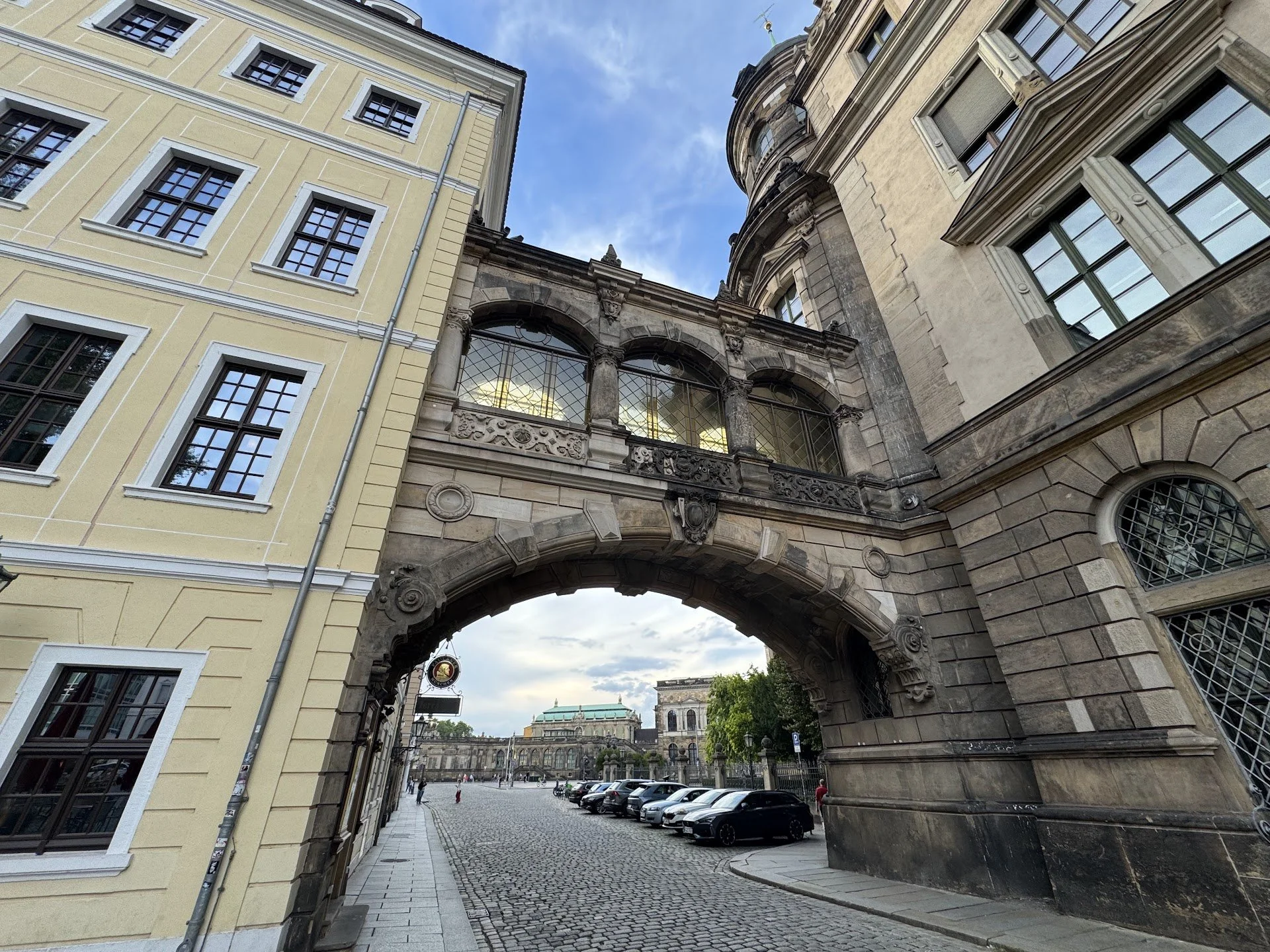 Street view showing historic buildings with an archway, cobblestone street, parked cars, and a cloudy sky.