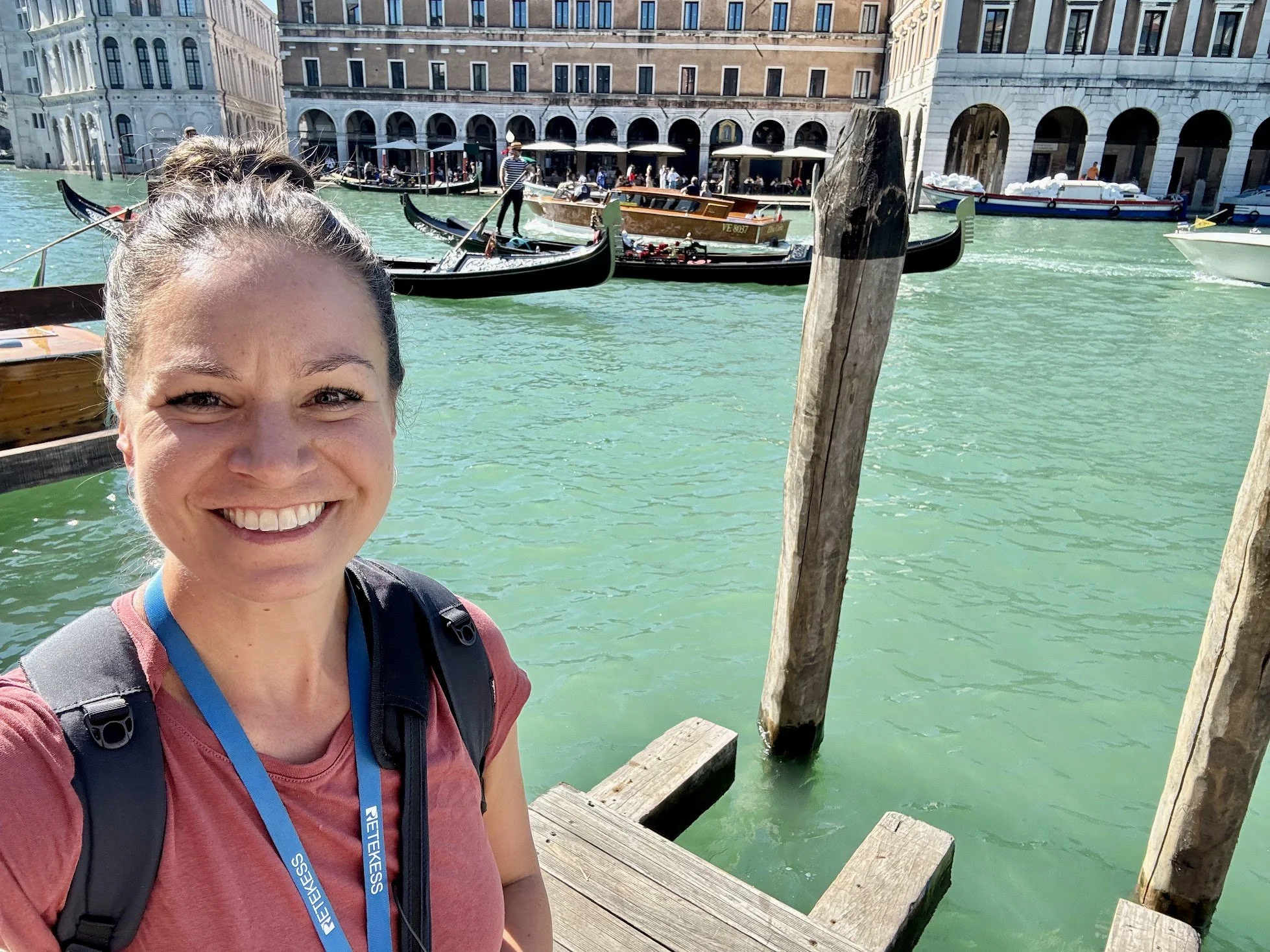 Smiling woman with a backpack taking a selfie on a dock in Venice, Italy, with gondolas and historic buildings surrounding the canal.