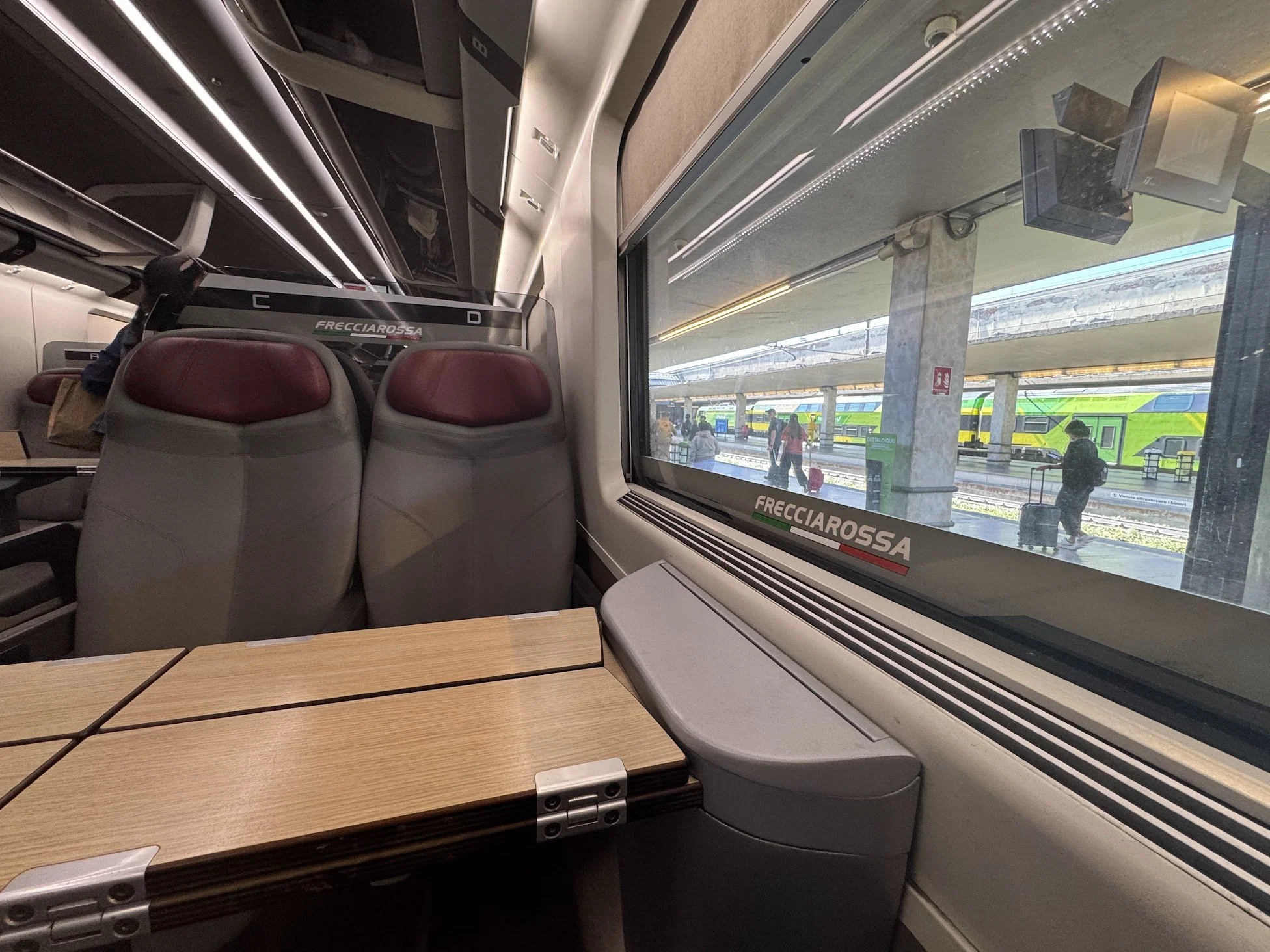 Inside a train compartment with red seats and a large window showing a train station platform with people walking and bags, with a green train outside.
