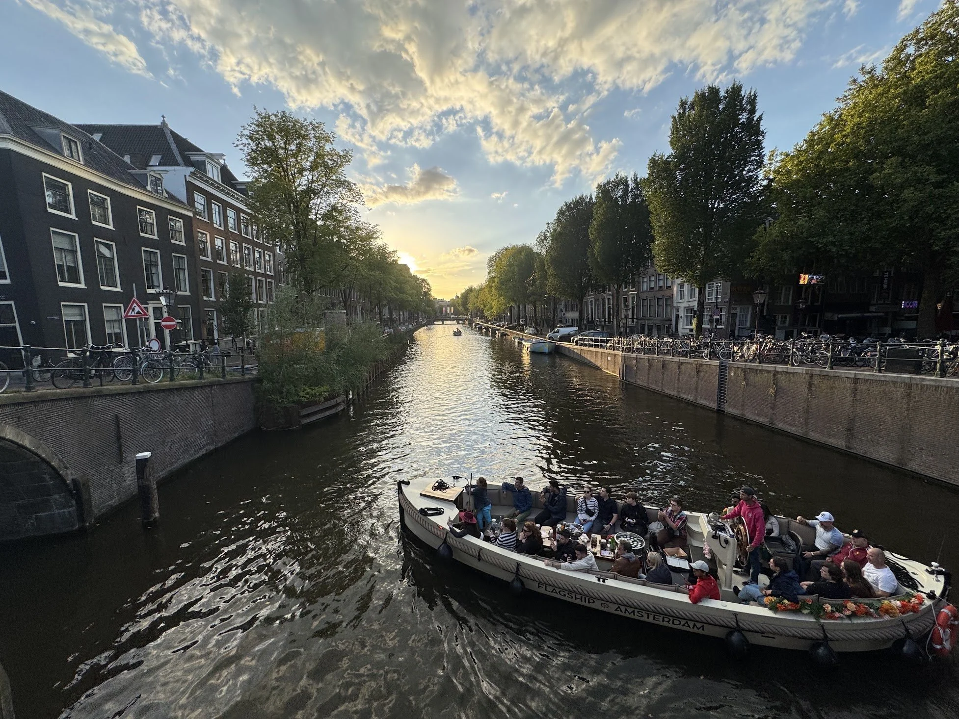 A boat with tourists sailing through a canal in Amsterdam, with historic buildings on either side and a sunset sky.