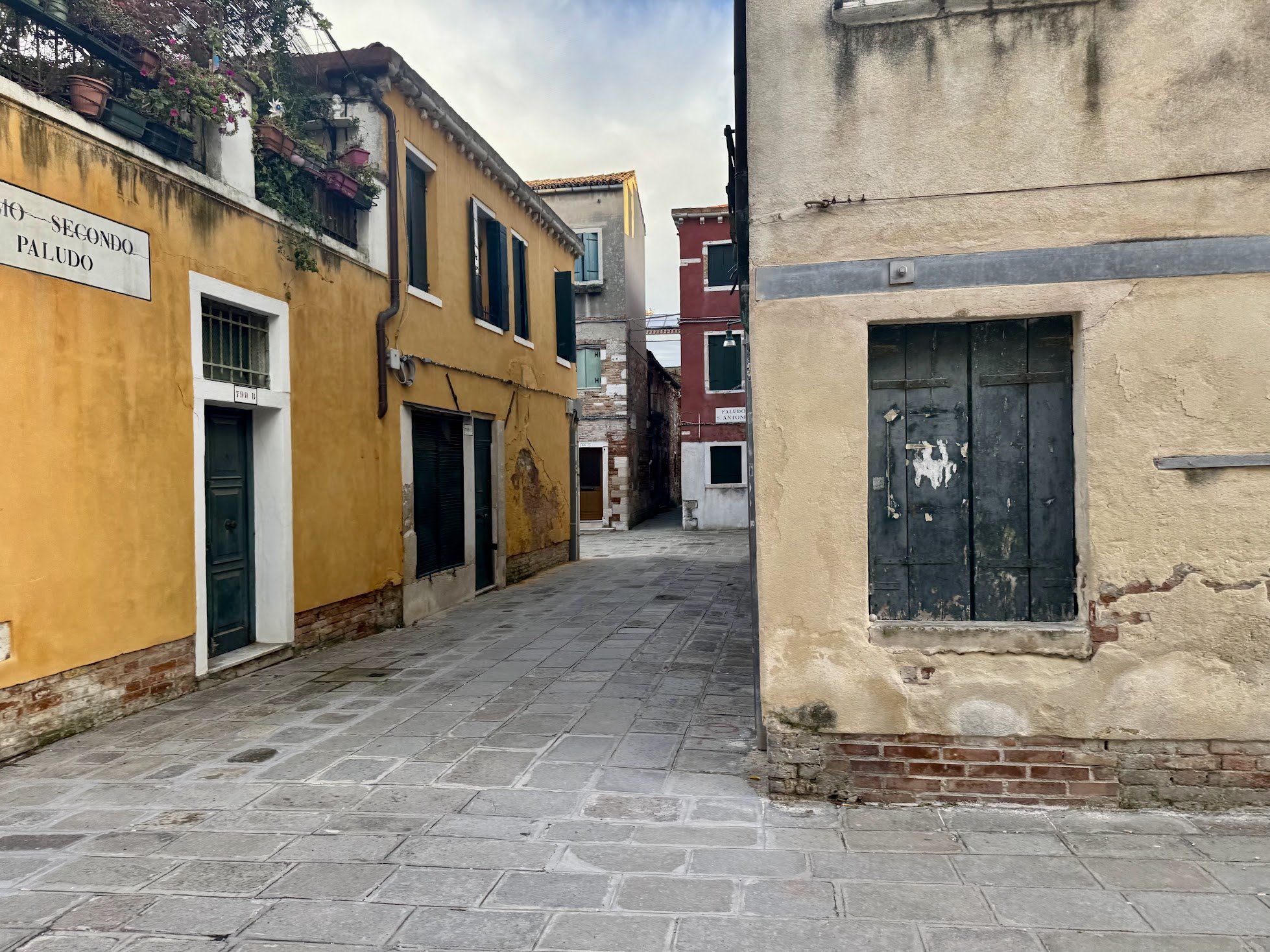A narrow, cobblestone street in an old European town, lined with colorful, weathered buildings with small windows and flowers on a balcony.