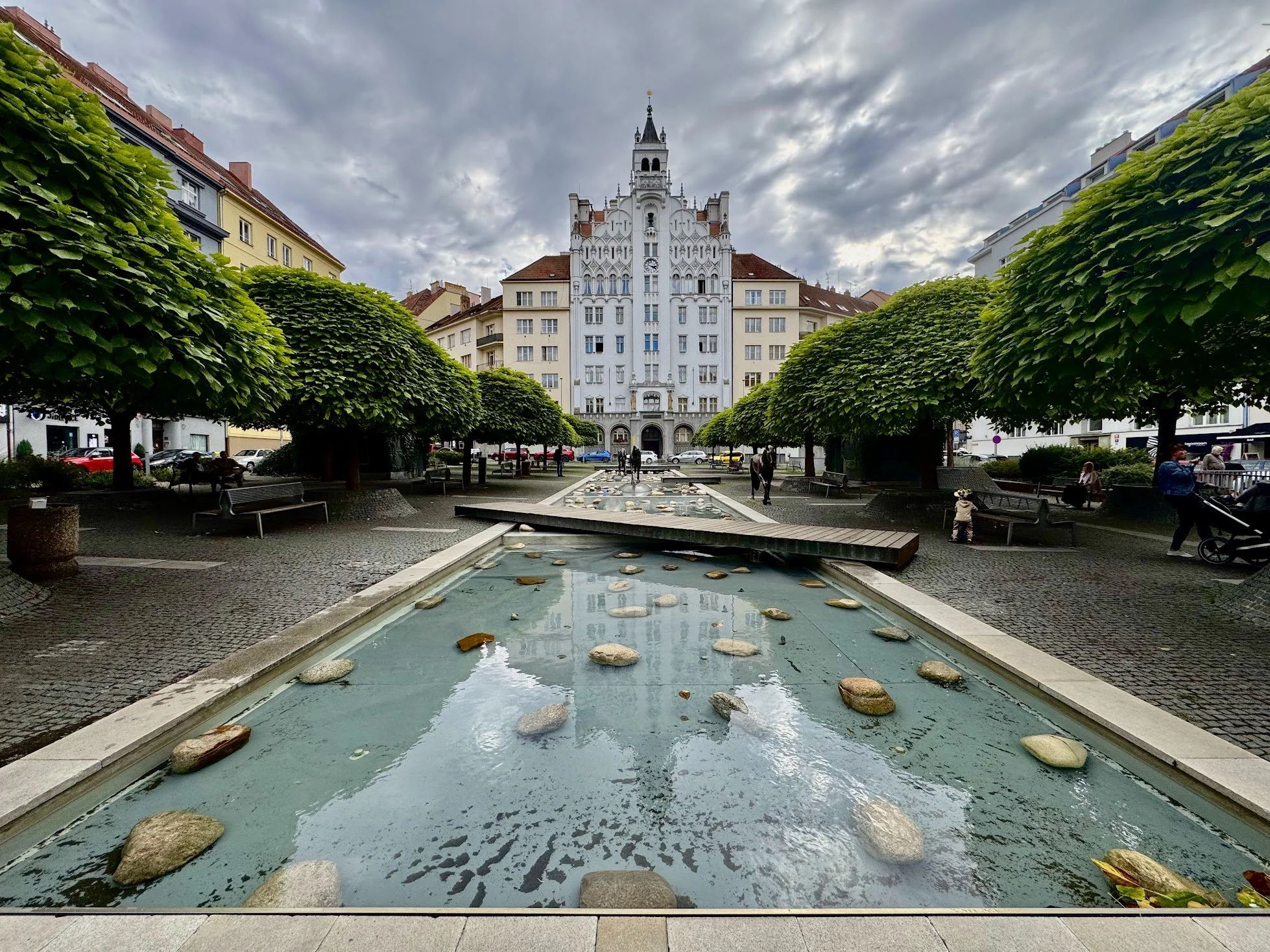 View of a city square with a water feature, surrounded by green trees, benches, and people walking. A large historic building with a tower is in the background under a cloudy sky.