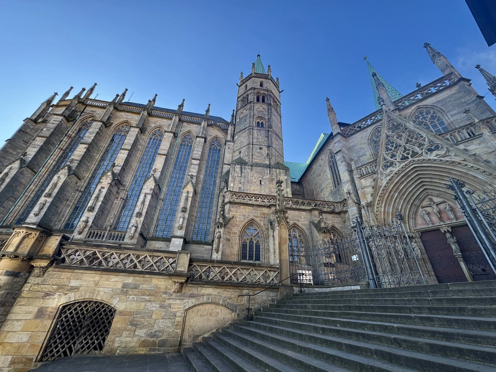 Photo of a historic Gothic church with tall spires, arched windows, and detailed stone carvings. The church has a stone exterior with green roofs, and steps leading up to the entrance, with a metal gate in front.