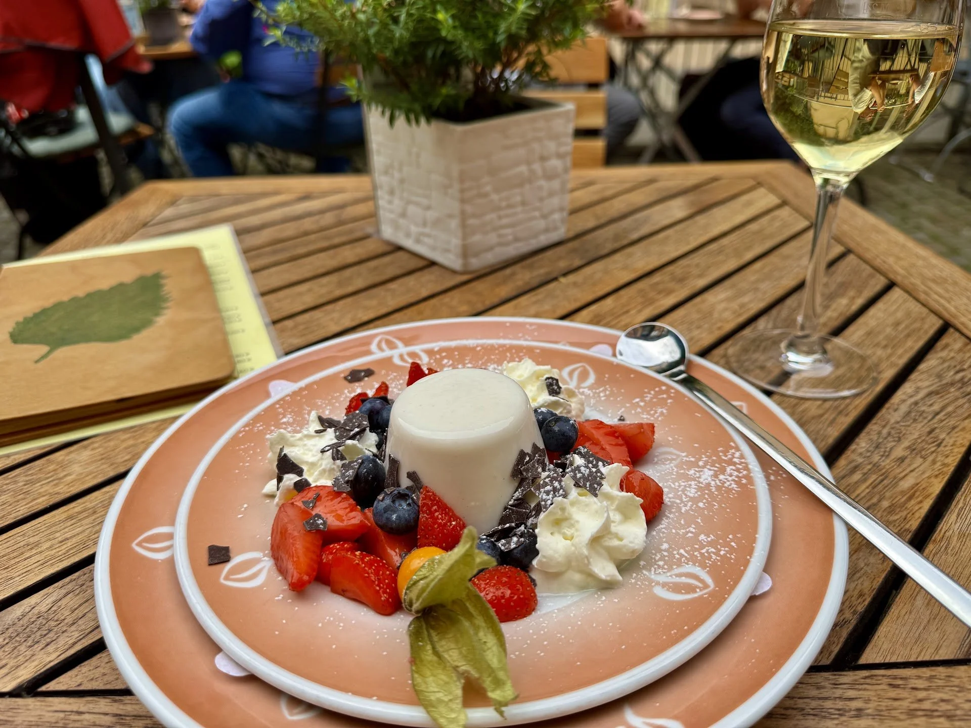 A dessert with strawberries, blueberries, chocolate shavings, whipped cream, and a white panna cotta on a peach-colored plate, on a wooden table. A glass of white wine and a floral centerpiece are visible in the background.