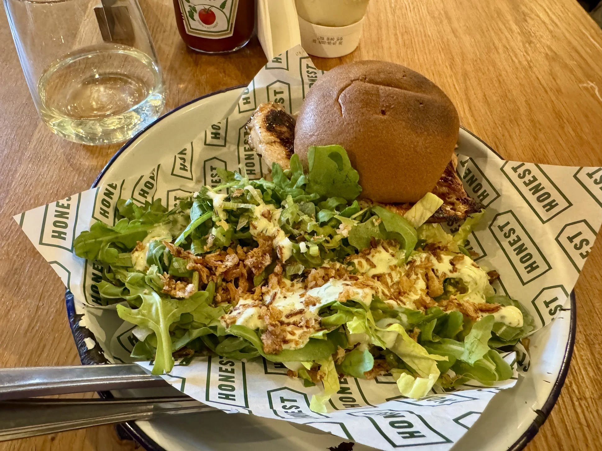 A plate of food featuring a sandwich with a bun, leafy greens, and a chicken or fish fillet, served with potato chips and a side of salad. There is a glass of water and condiments, including ketchup and a small container of mayonnaise, on a wooden ta