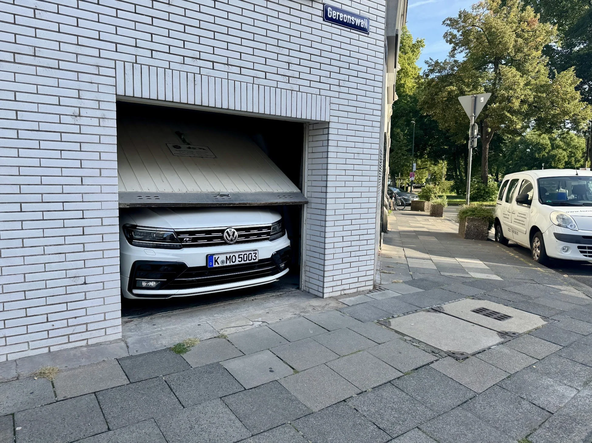 White brick building with a parking garage entrance, a Volkswagen car inside, and a street with parked cars and trees on a sunny day.