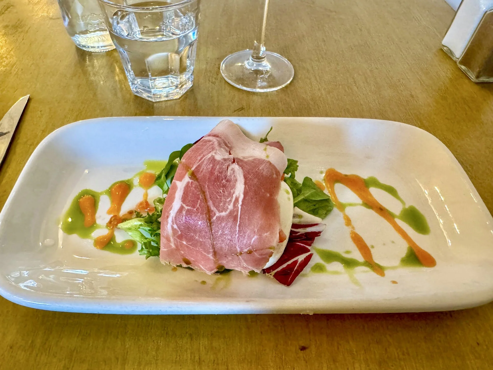 A white rectangular plate with a serving of prosciutto wrapped around a green leafy salad, garnished with colorful sauces, on a wooden table. In the background, there is a glass of water and an empty wine glass.