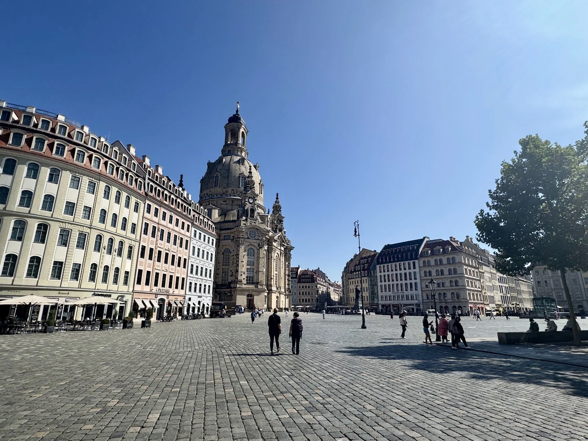 A European city square with cobblestone paving, historic buildings, and a large church with a domed tower. People are walking and sitting in outdoor cafes on a sunny day, with a tree visible on the right side.