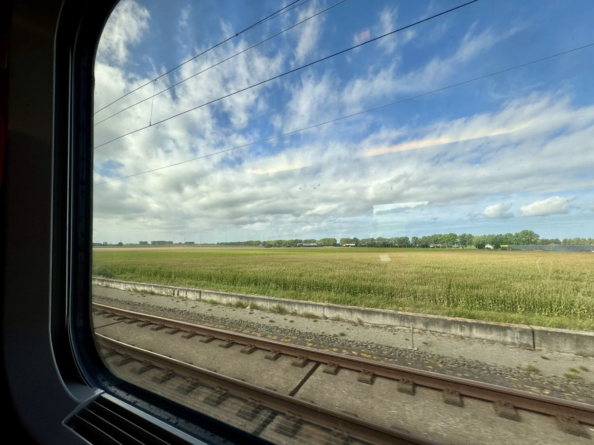 View of green fields, blue sky with scattered clouds, and train tracks from inside a train window.