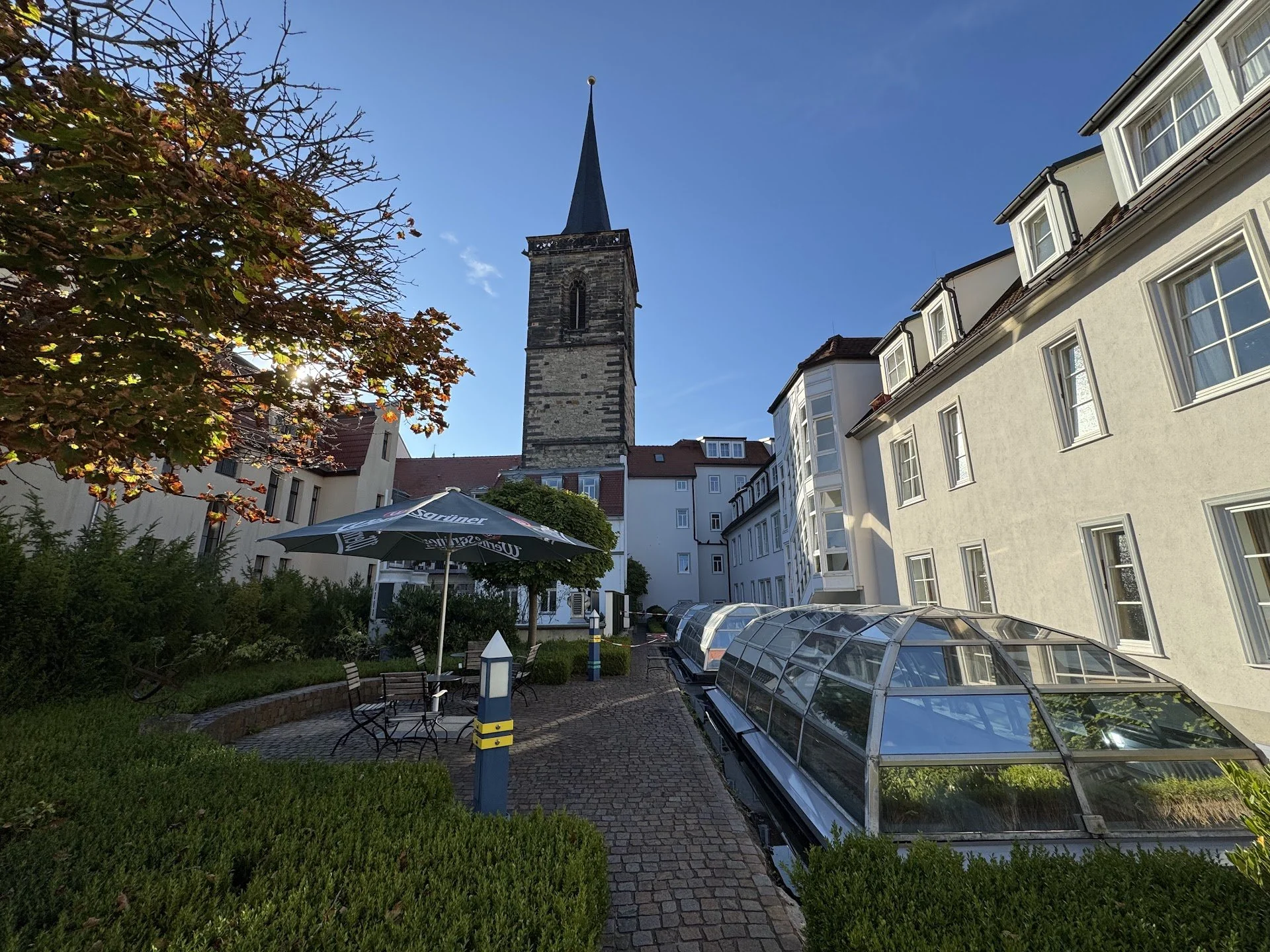 Outdoor garden area with benches, tables, an umbrella, and glass-roofed structures, bordered by bushes and surrounded by residential buildings and a church with a tall steeple.