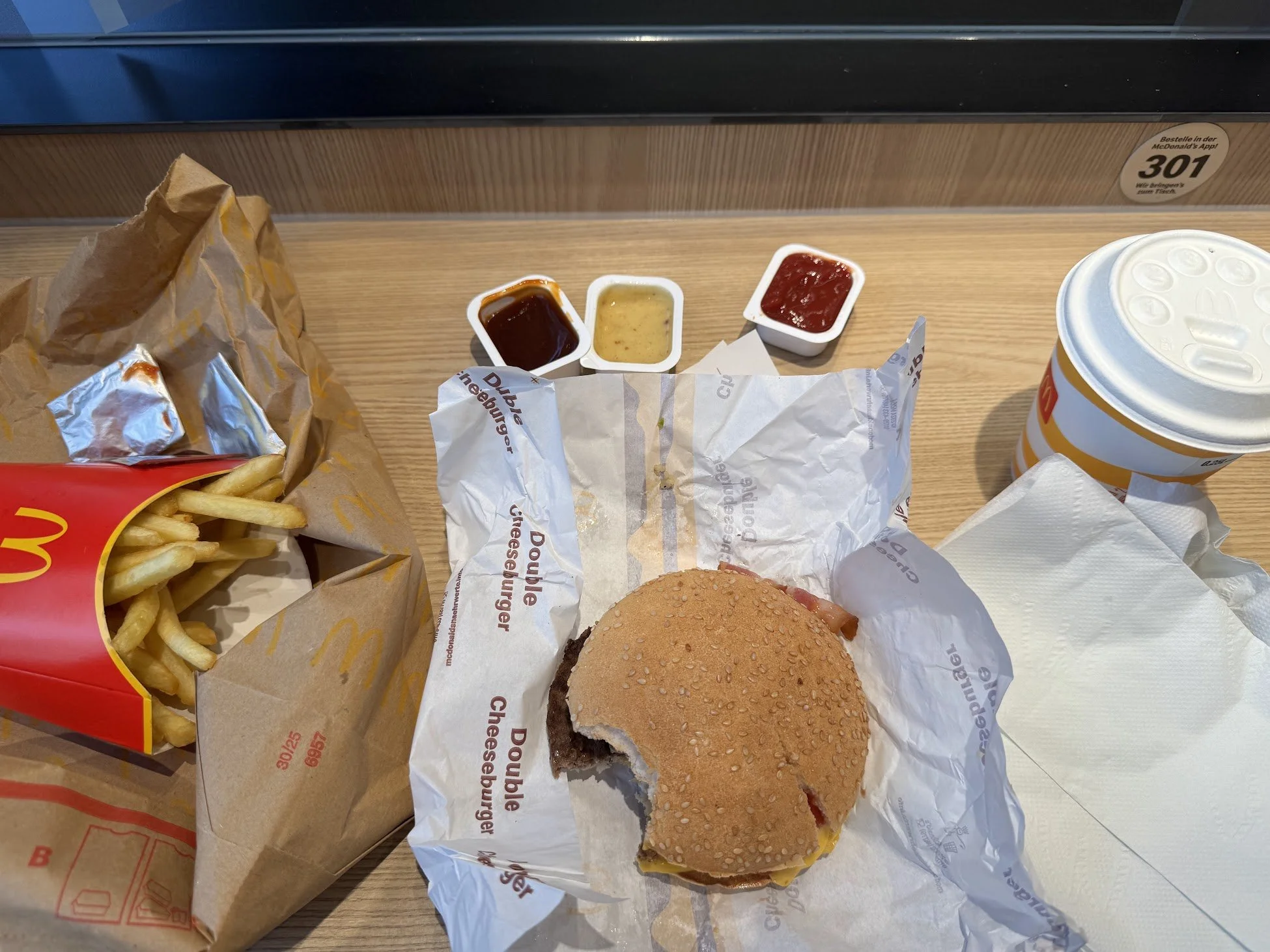 Fast food meal on a tray including a cheeseburger in a sesame seed bun, a bag of French fries, and three small containers of dipping sauces—barbecue, honey mustard, and ketchup.