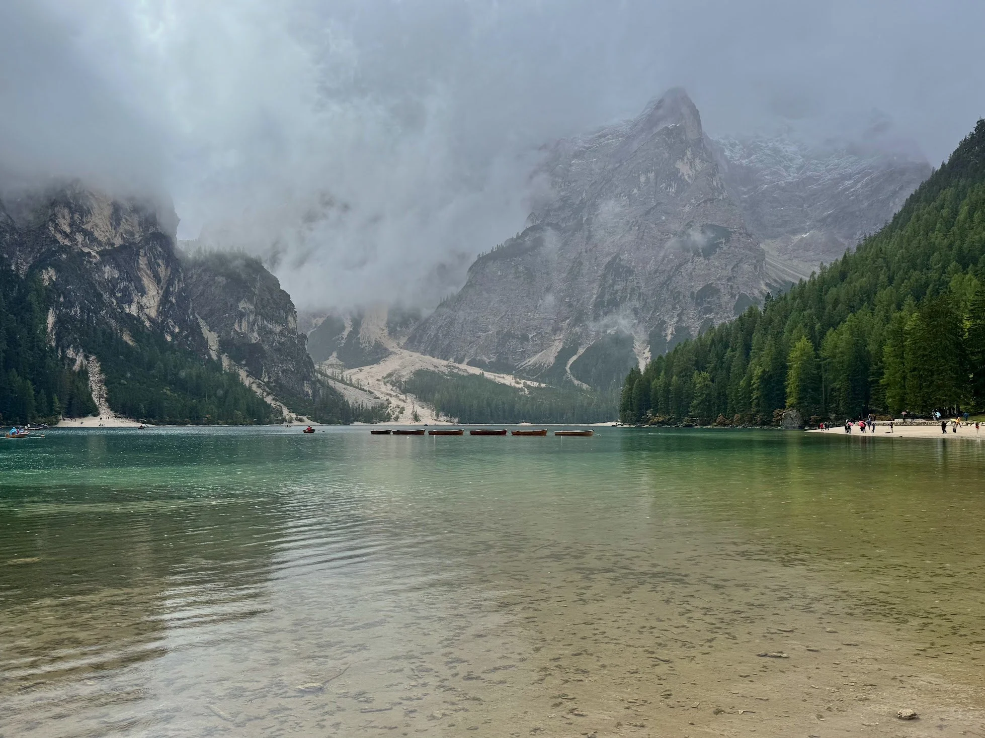 A scenic mountain landscape with a foggy sky, tall mountain peaks, dense green pine forests, a serene lake with small boats, and a sandy beach with people walking.
