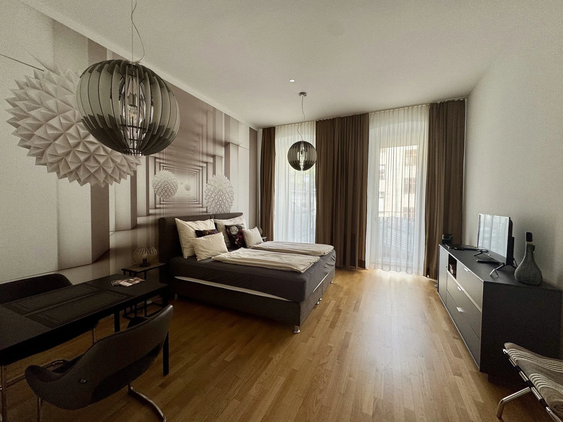 A living room with large windows covered by sheer and opaque curtains. There are two modern pendant lights hanging from the ceiling. A sofa with pillows is positioned along the wall, and a decorated wall art is behind it. A TV is on a white console t