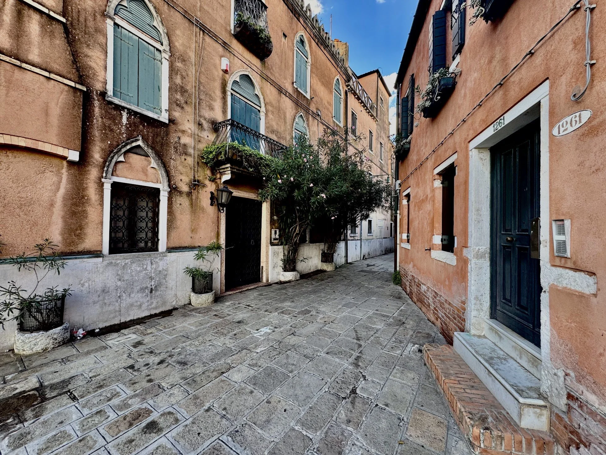 A narrow European street with old, colorful buildings on both sides, featuring arched windows with shutters, decorative balconies with plants, and a cobblestone pavement.
