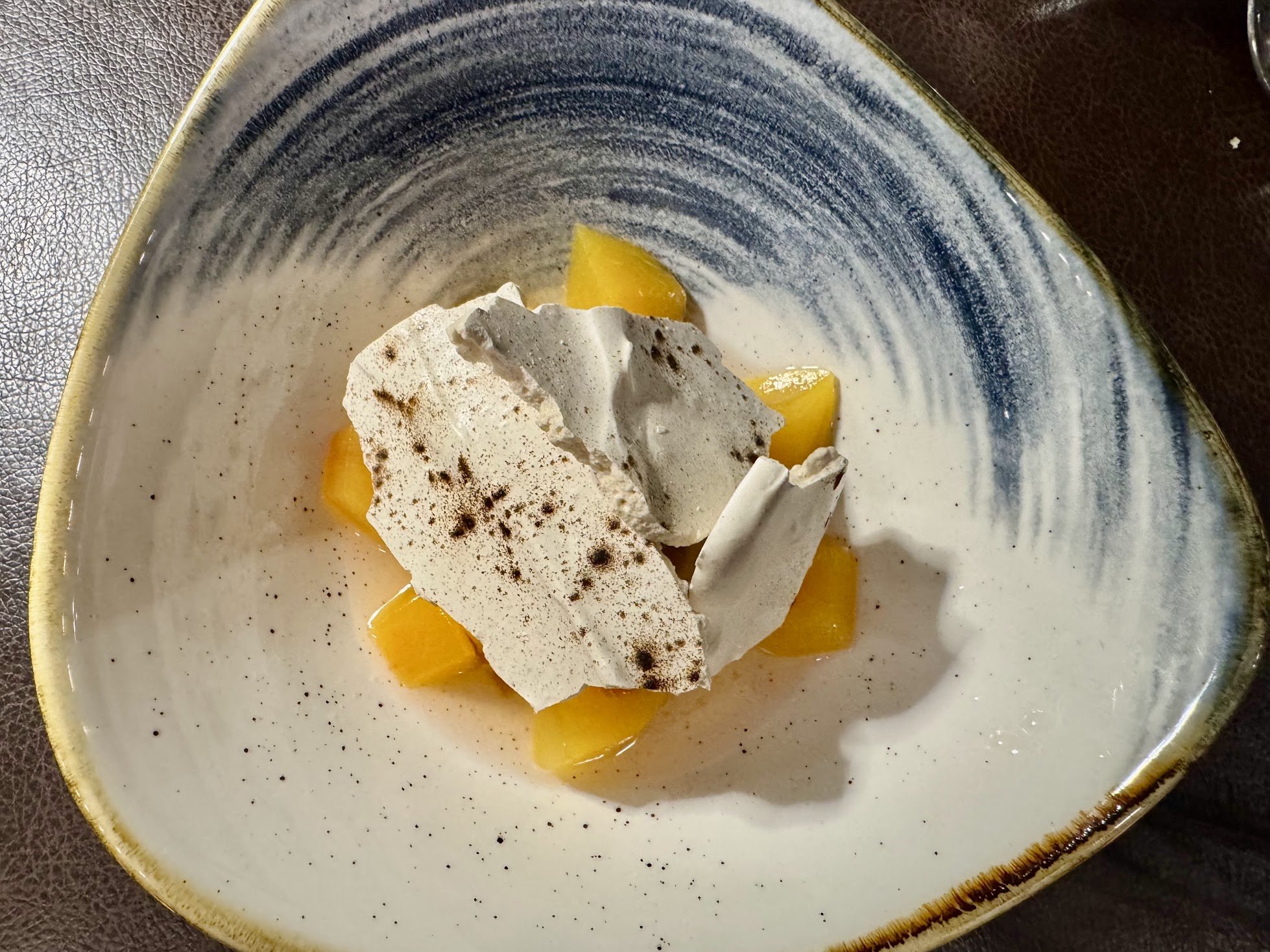 A dessert with a dollop of whipped cream sprinkled with cocoa powder, garnished with slices of yellow fruit in a decorative bowl with a gradient blue and white glaze.