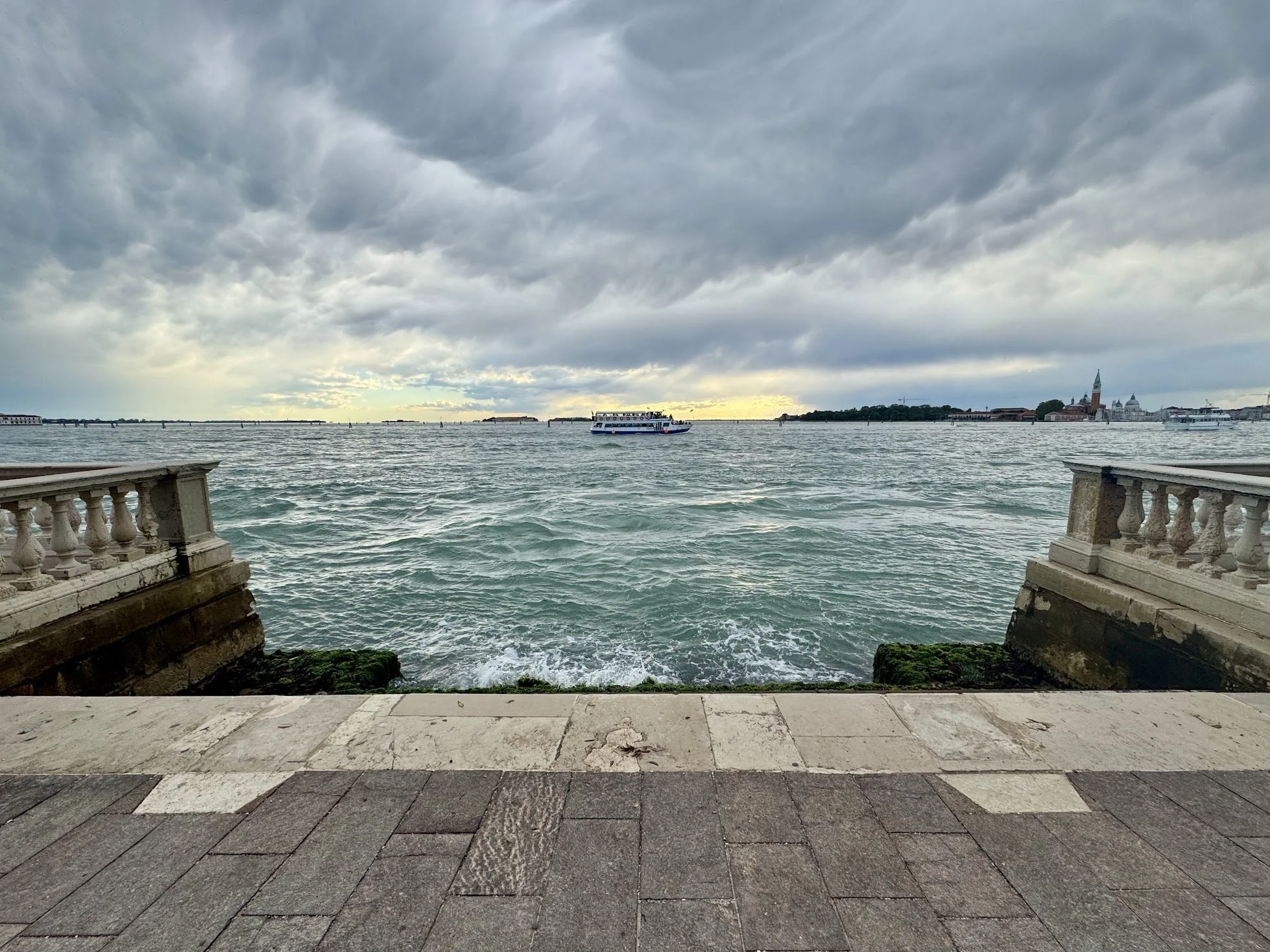 View of the water with a boat and a city skyline in the background, seen from a stone-lined waterfront promenade with a stone railing.