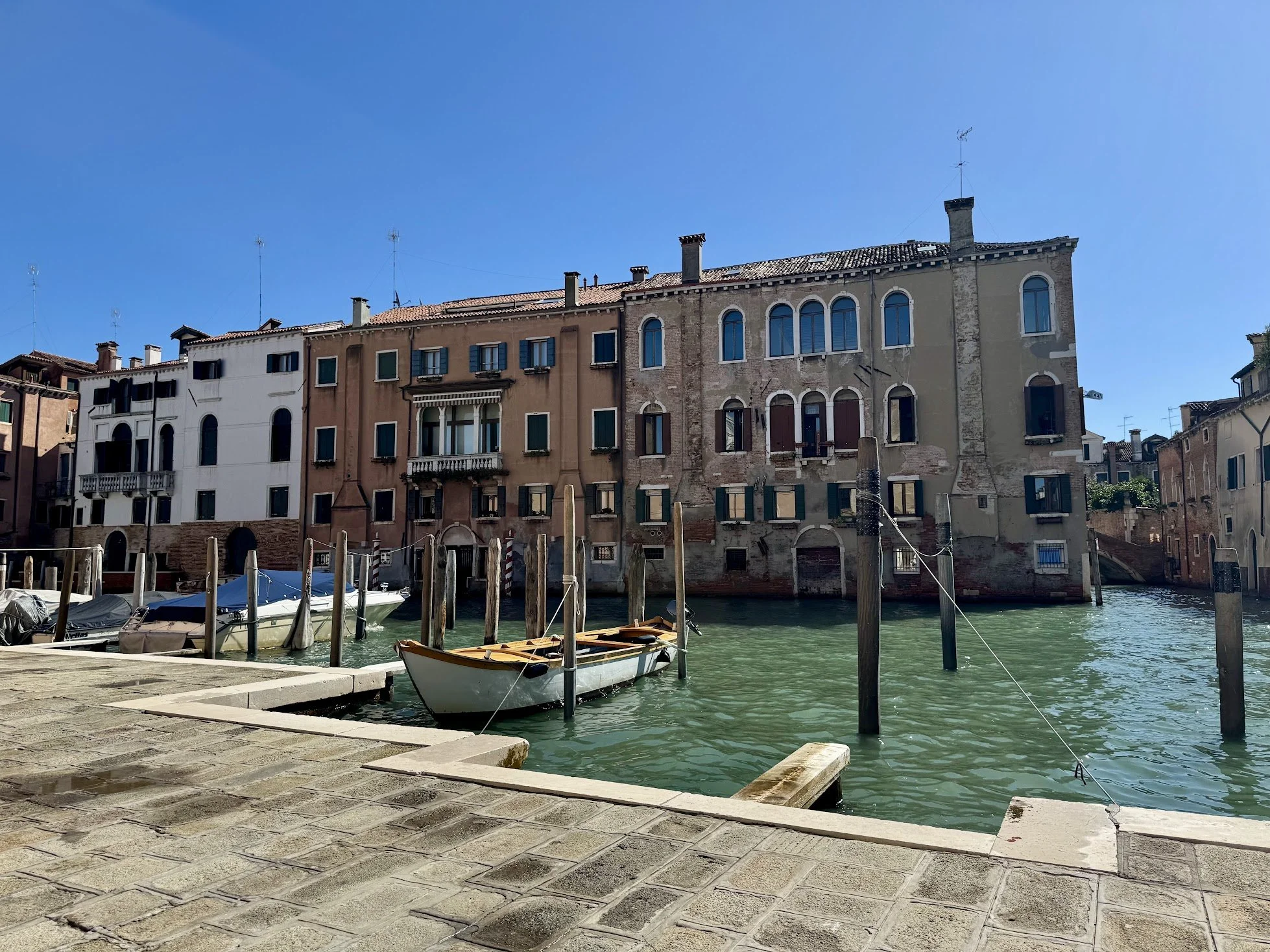 Colorful historic buildings along a canal in Venice, Italy, with boats docked at the water's edge under a clear blue sky.