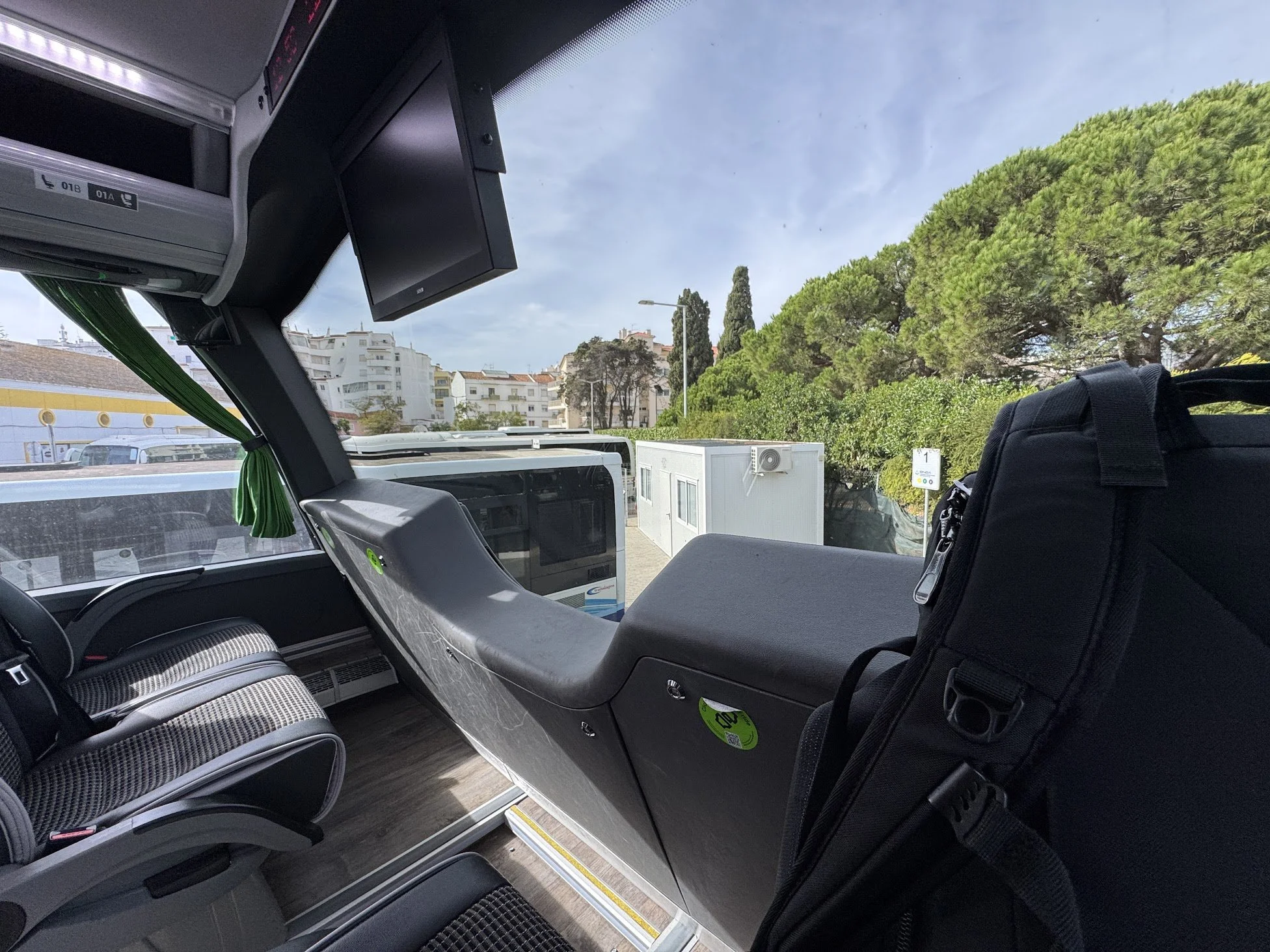 Interior of a bus with two rows of gray and black patterned seats, a black luggage compartment and a monitor screen. Outside view through the bus window shows trees and buildings under a partly cloudy sky.
