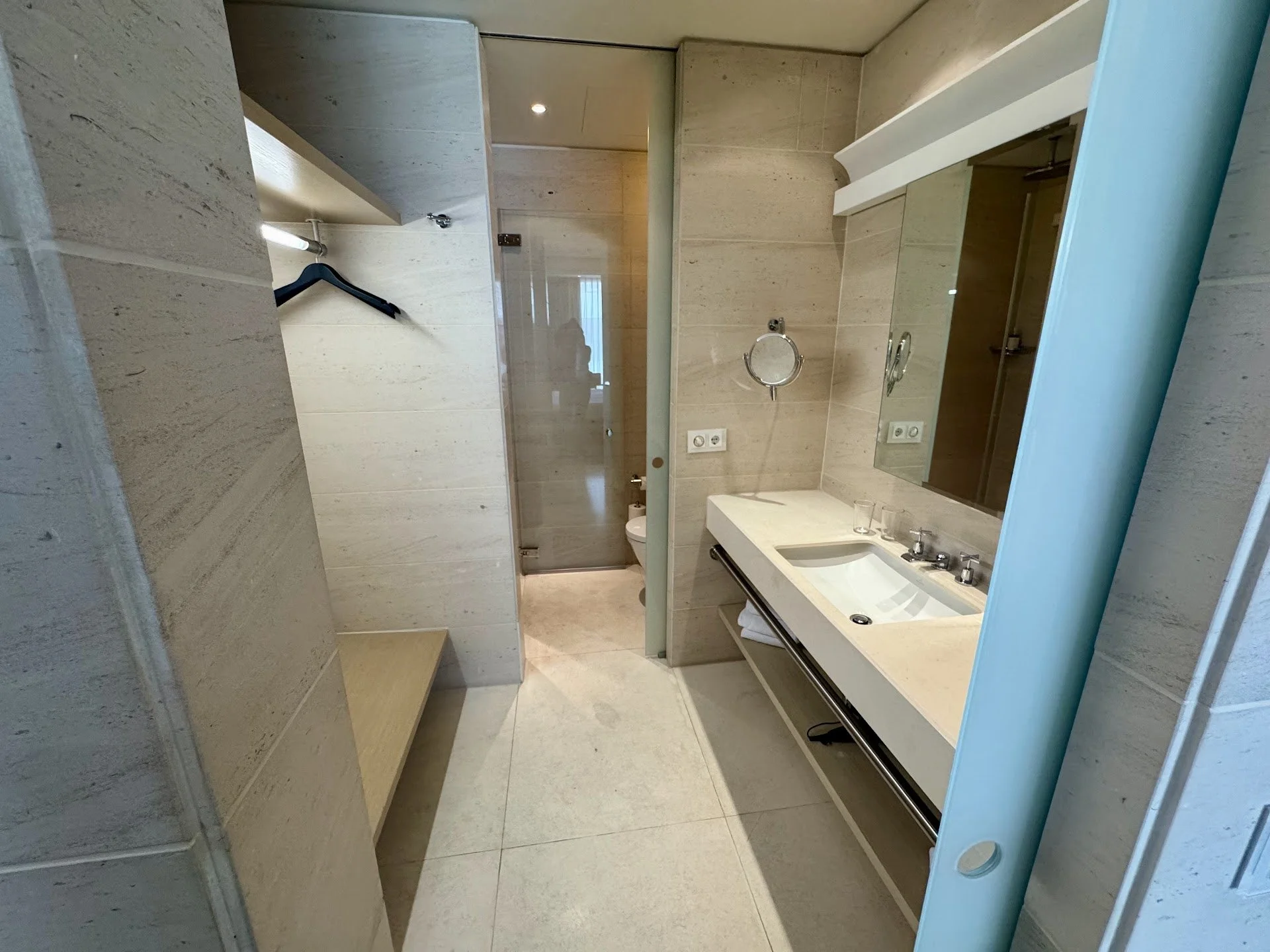 A modern hotel bathroom with a beige tiled wall, a white sink with a black drawer, a mirror, a soap dispenser, a glass, a white towel, a mirrored cabinet, a hairdryer, and a shower stall with a glass door.
