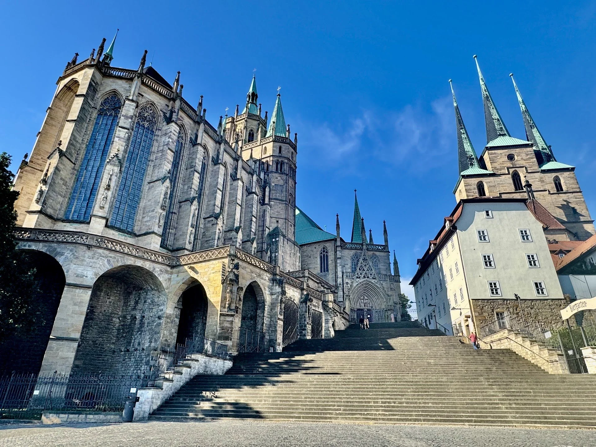 A historic Gothic-style church with tall spires and large stained glass windows, situated at the top of a wide staircase in a European town, under a clear blue sky.