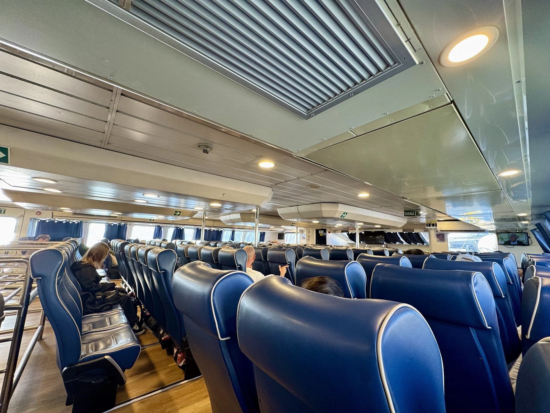 Interior of a ferry or airplane with rows of blue seats, passengers sitting and looking at devices, large windows with curtains, ceiling with air vents and lights, signage including an exit sign and a 'Zona B' sign.