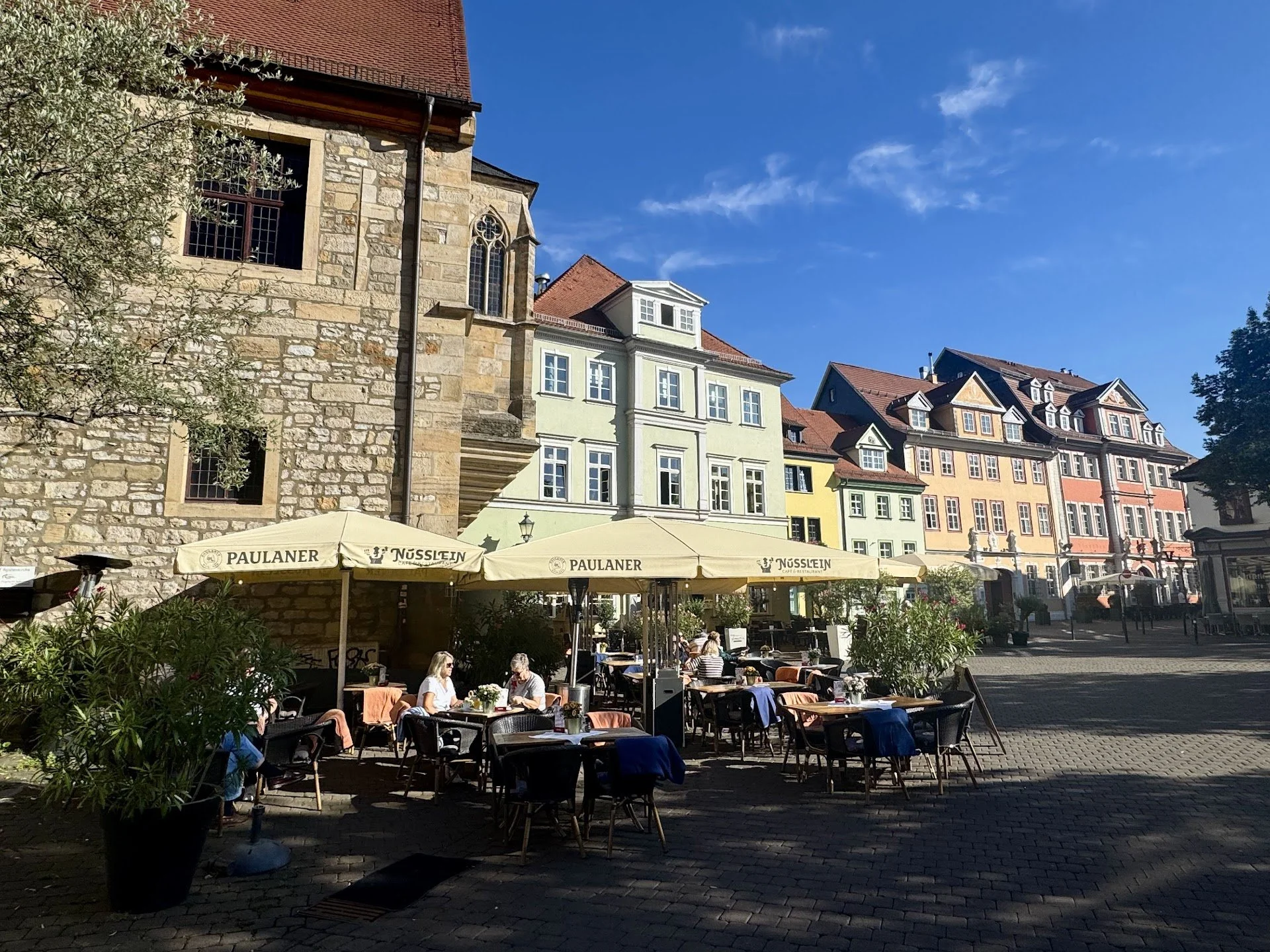 Outdoor cafe with yellow umbrellas and people sitting at tables, surrounded by colorful historic buildings and a stone church in the background under a bright blue sky.