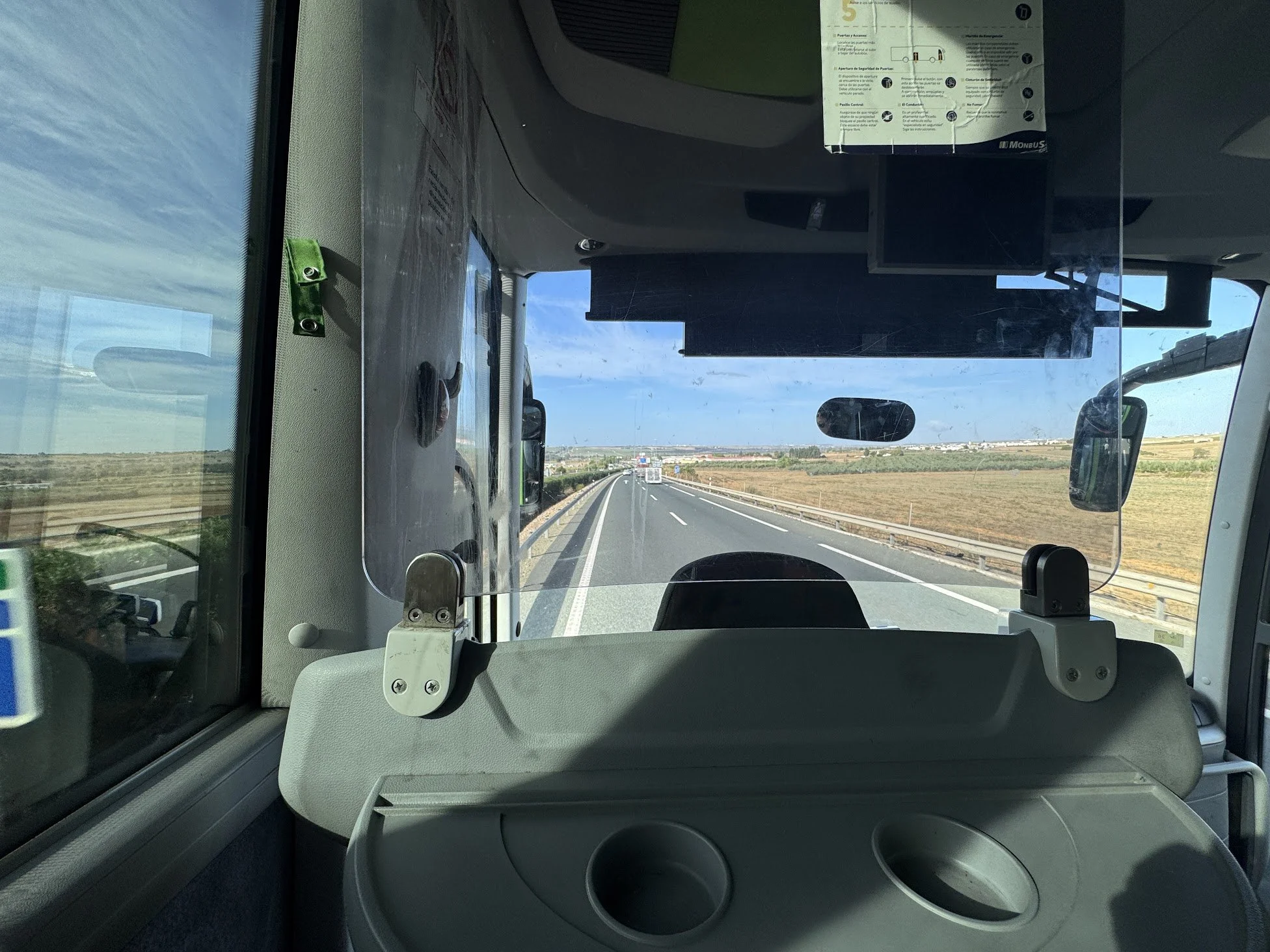 Looking through the front window of a bus, showing the road ahead with vehicles, landscape, and blue sky with clouds.