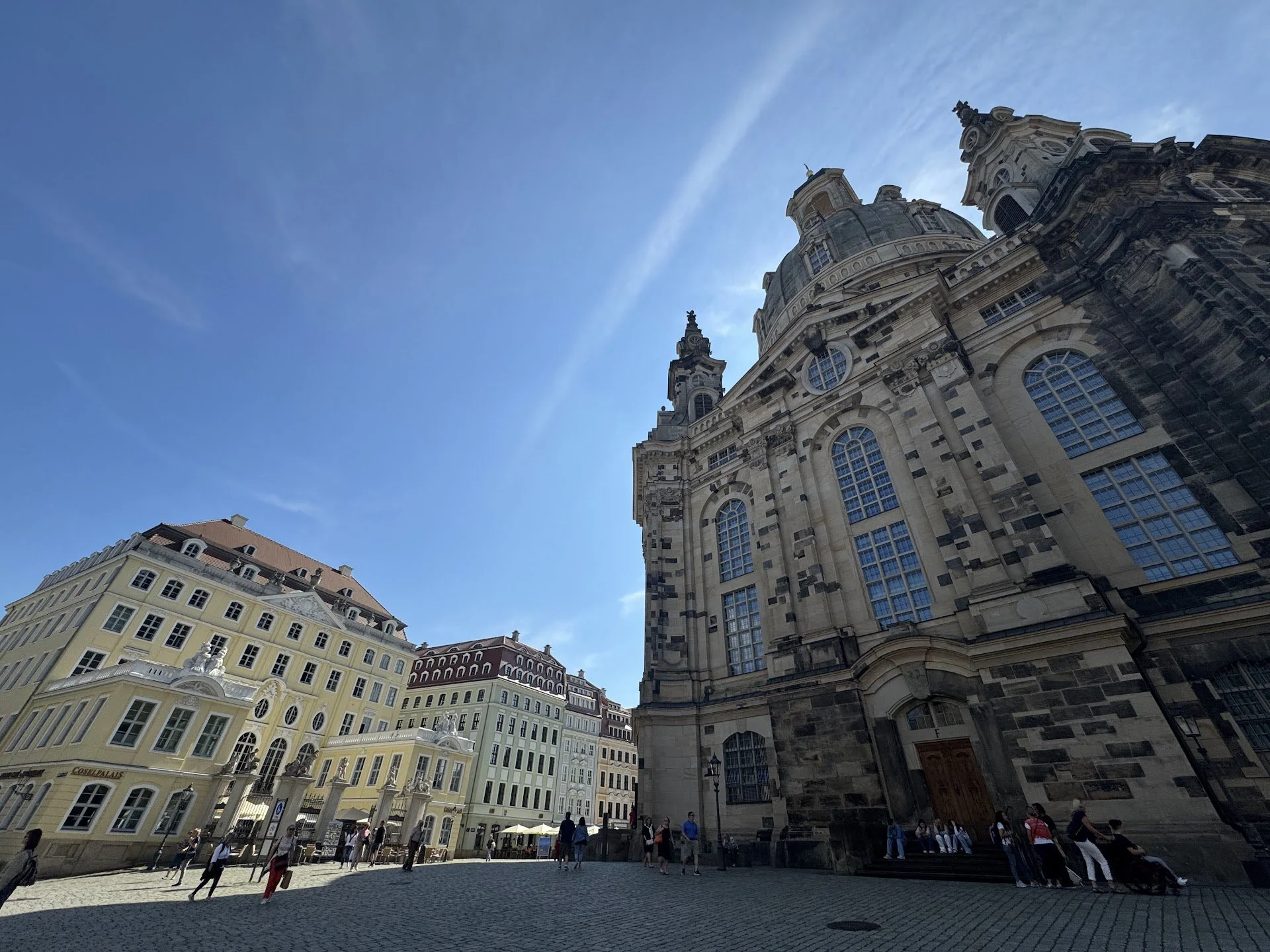 A large historic church with intricate stonework and domes, surrounded by colorful buildings, with people walking and sitting outside under a blue sky.
