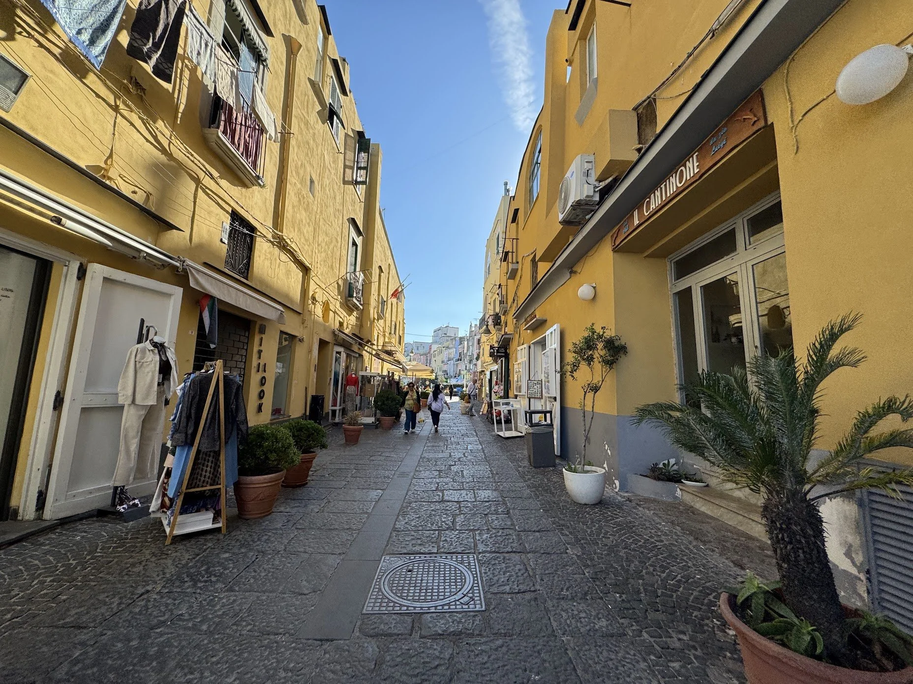 A pedestrian street lined with yellow buildings, outdoor shops, and potted plants, with people walking and browsing on a sunny day.