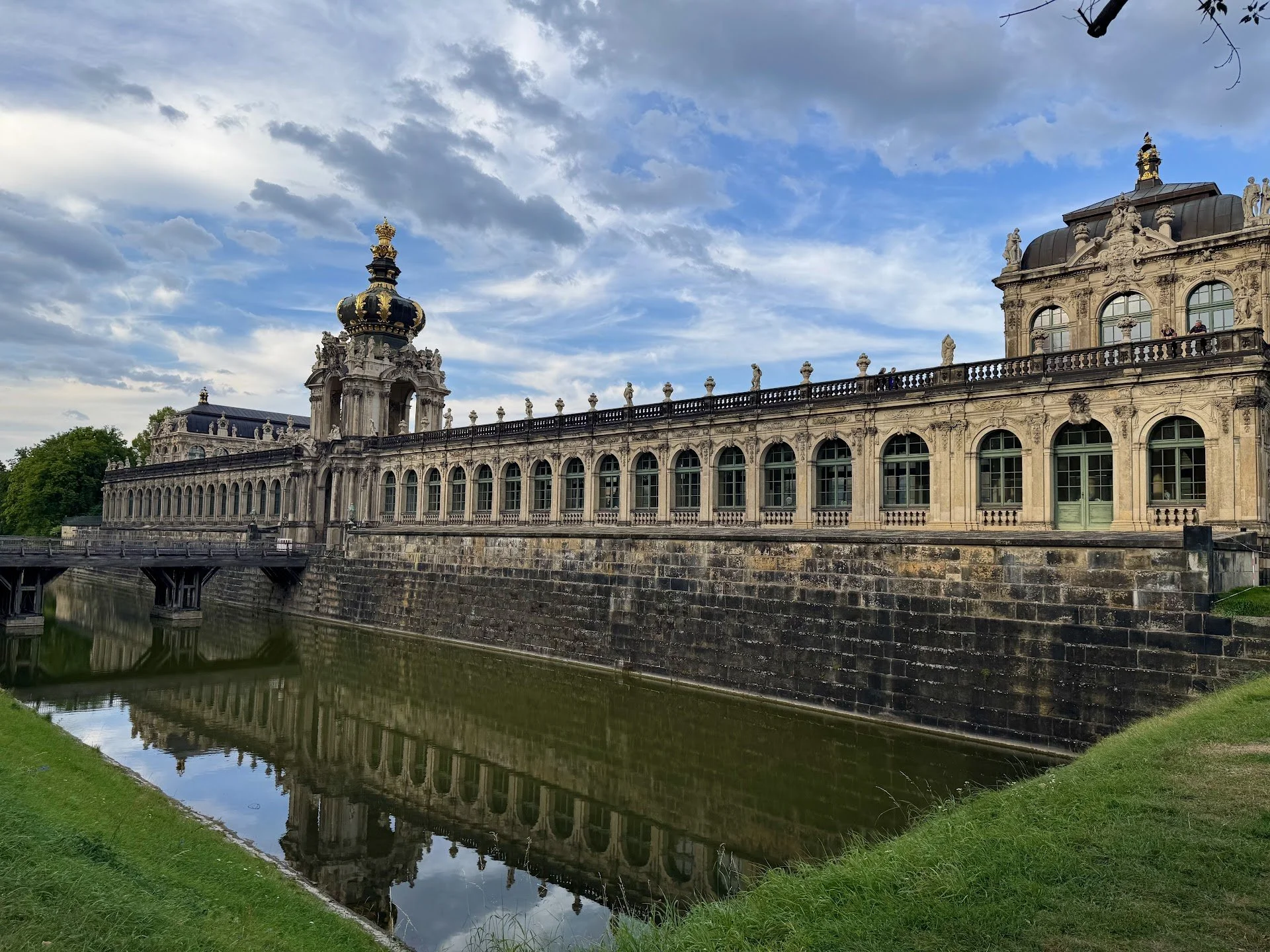 A historic Baroque-style building with intricate carvings and statues, reflected in the river below, under a partly cloudy sky.