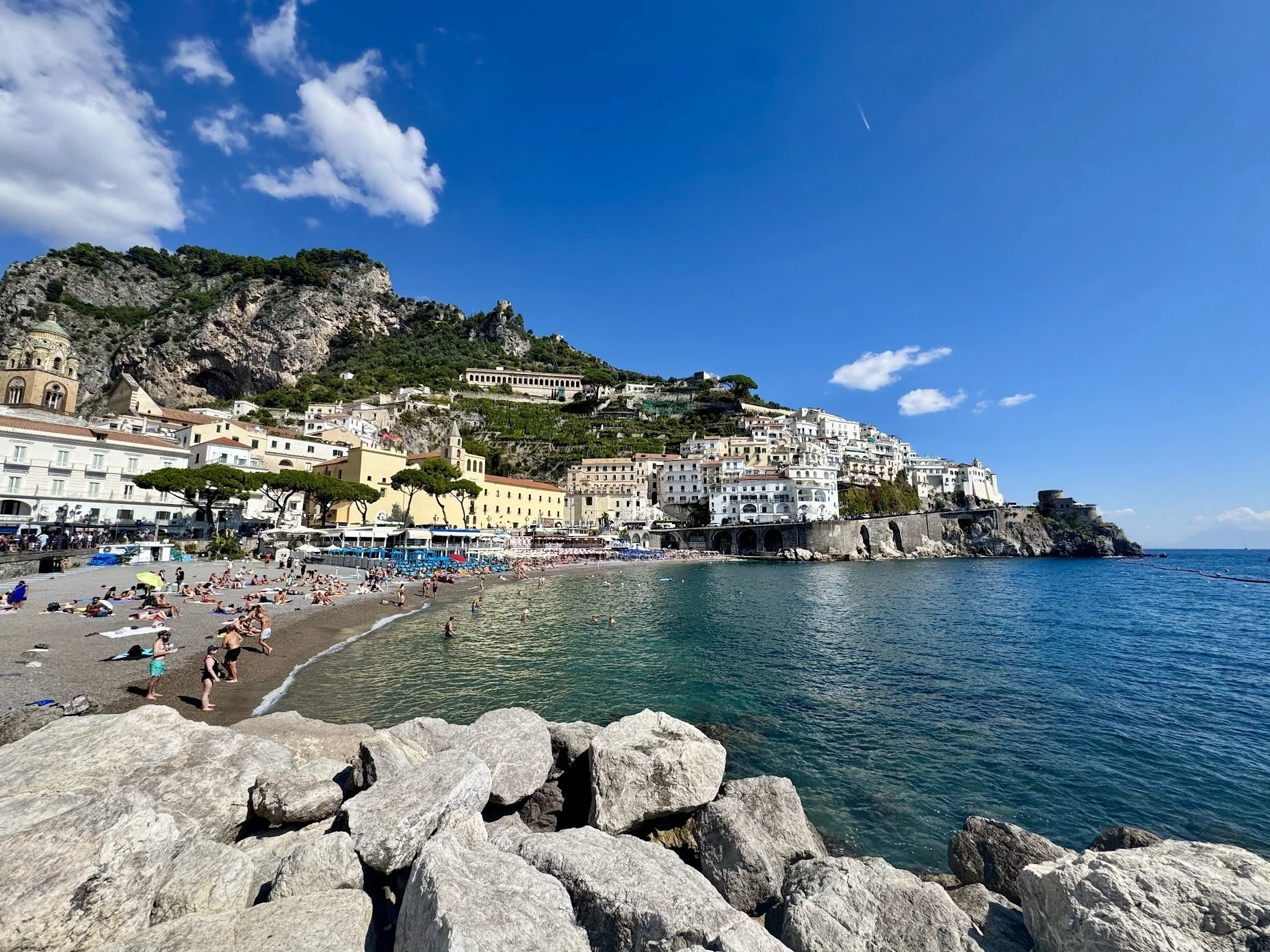 A beach with many people relaxing on the sand and swimming in the water, with a hillside town featuring colorful buildings and a mountain behind it, under a clear blue sky.