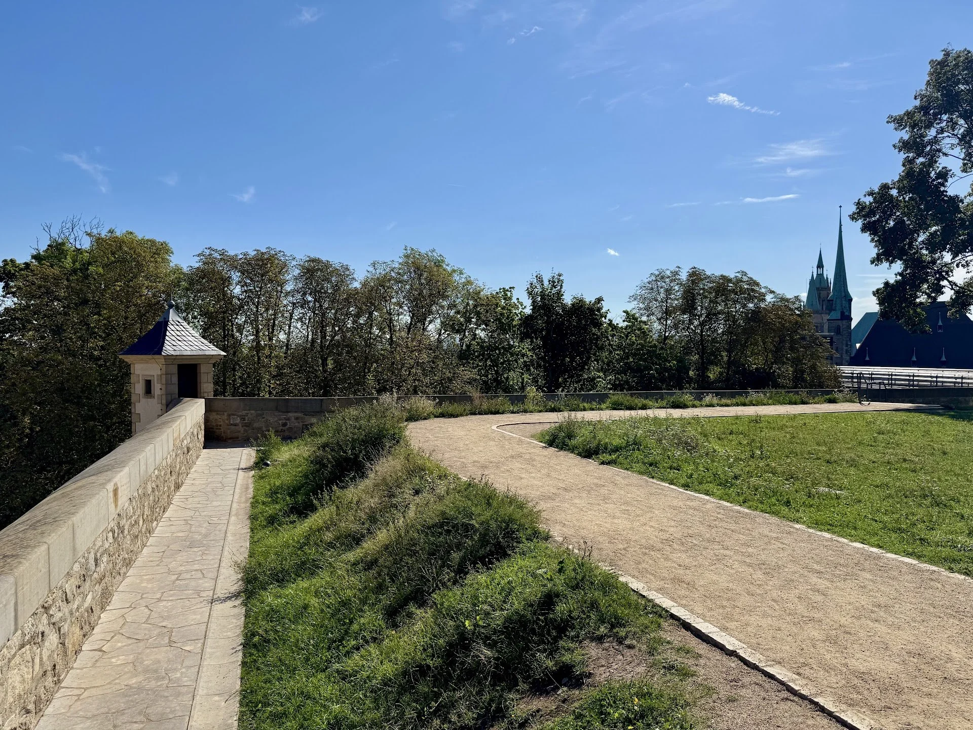 A park path with a stone wall on the left and grass on the right, leading towards a cityscape with church steeples in the distance and trees lining the horizon under a clear blue sky.