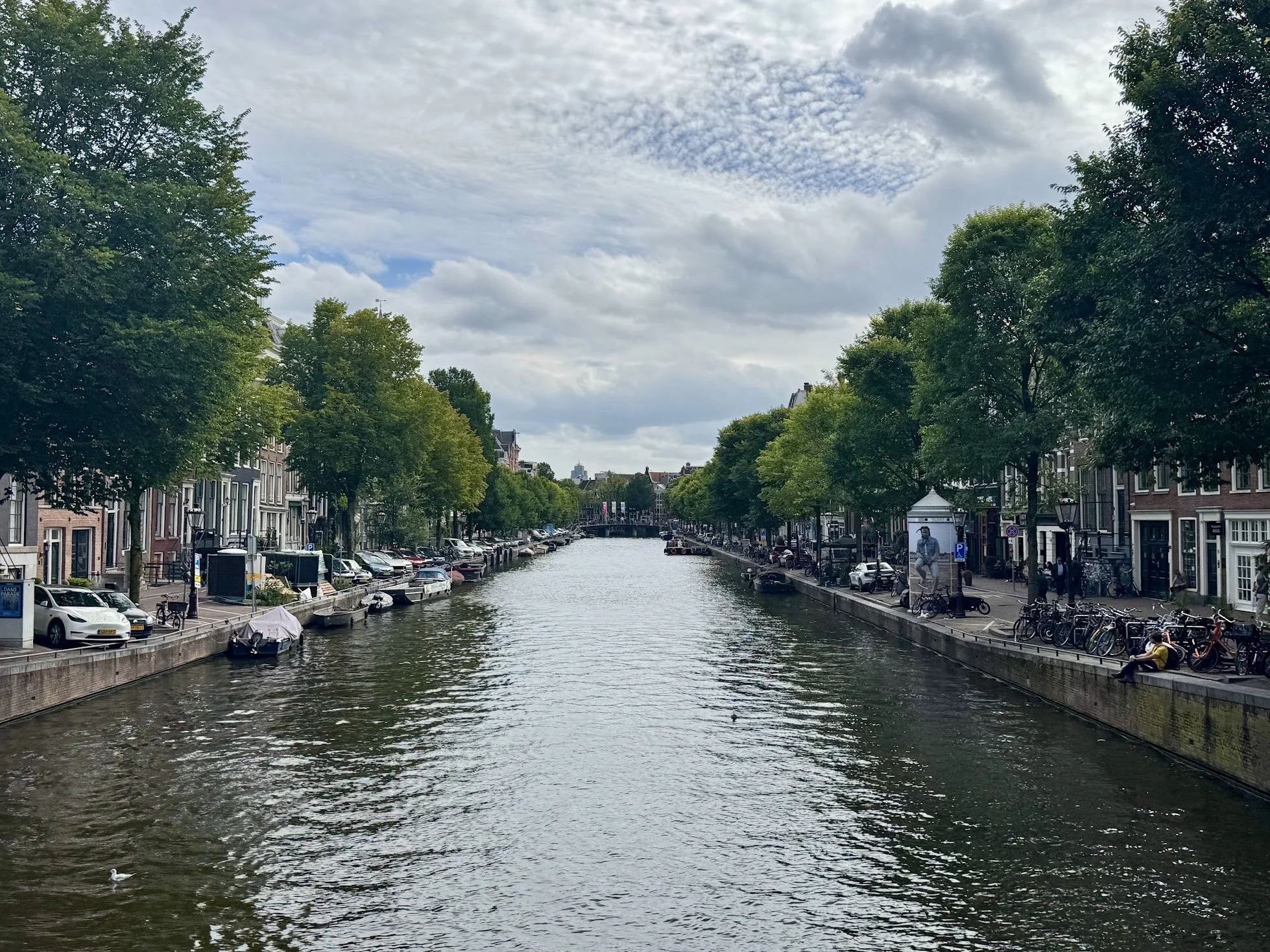 A canal in a city with trees lining the sides and buildings visible in the background. There are boats in the water and cars parked along the street. The sky is partly cloudy.
