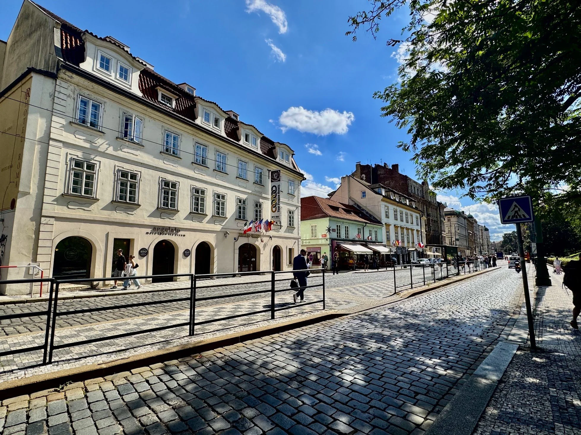 A street scene with cobblestone pavement, multi-story buildings with shops and flags, and a group of people walking on a partly shaded sidewalk under a bright blue sky with scattered clouds.