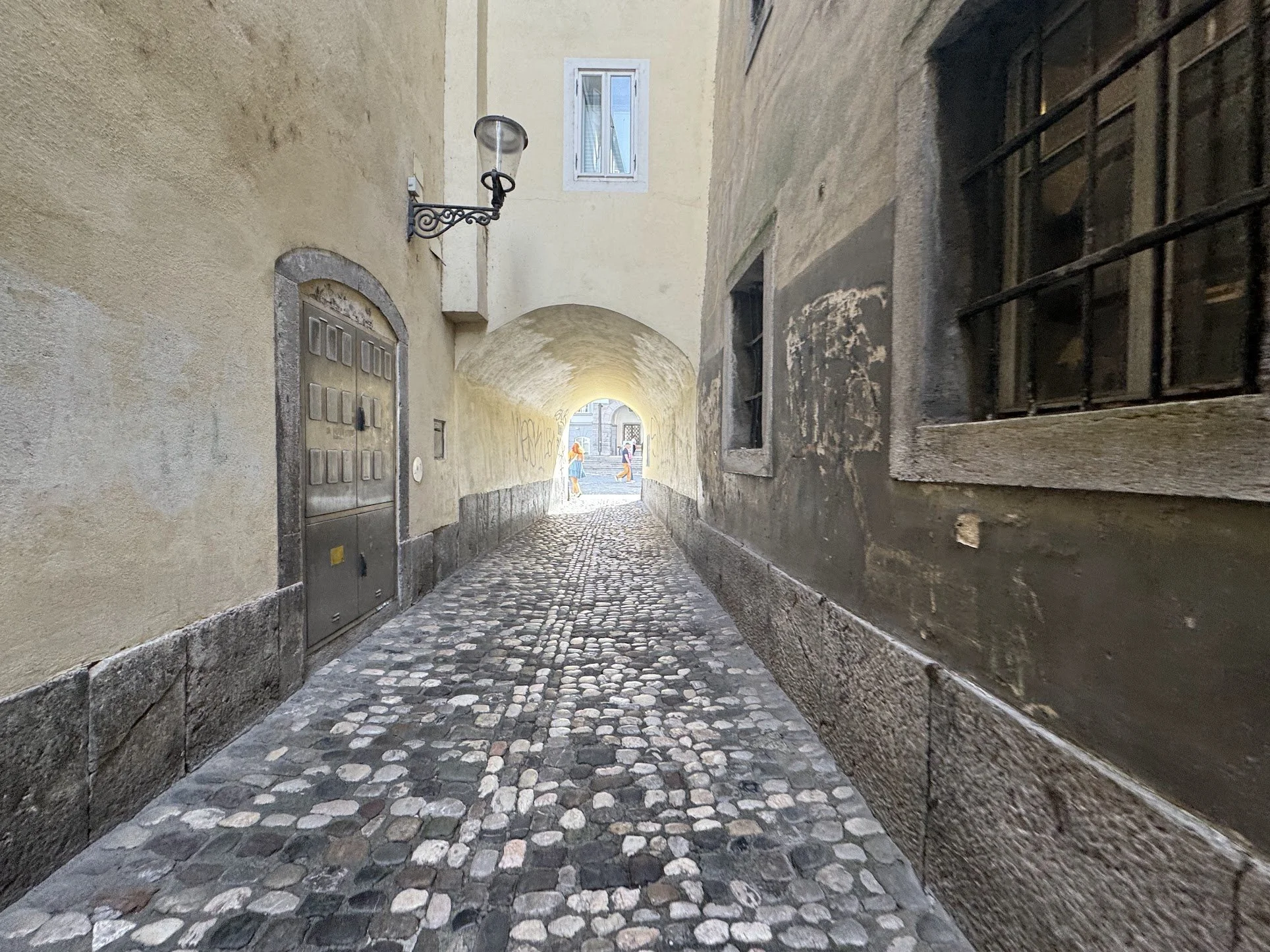 A narrow cobblestone alleyway with yellow and dark stone walls, an arched ceiling, and a small group of people walking in the distance.