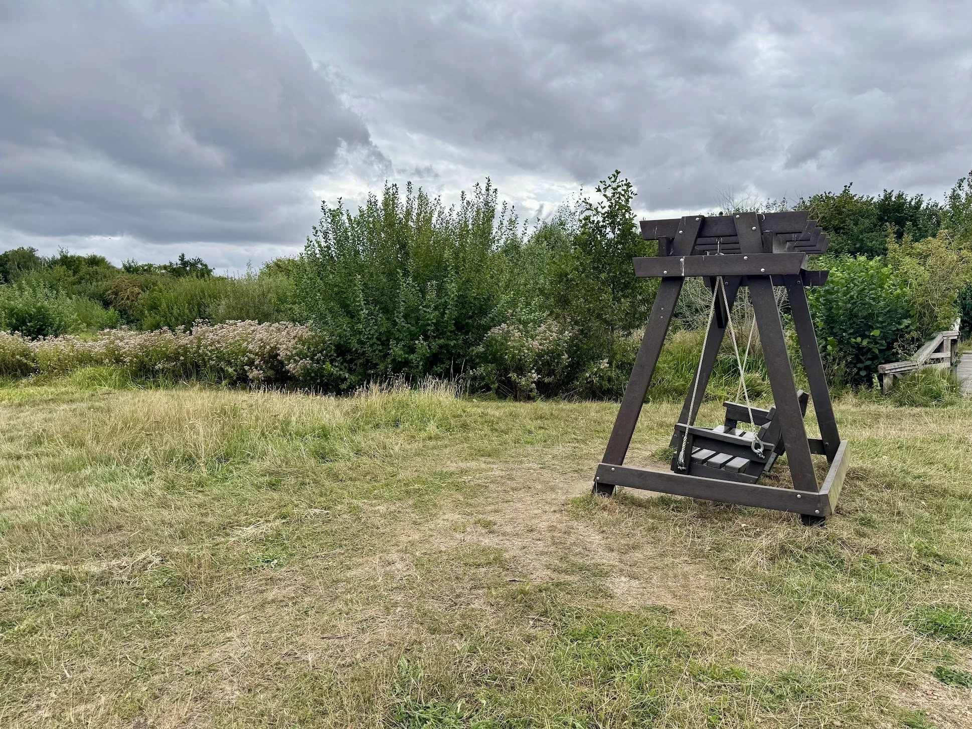 A wooden seesaw swing in a grassy field with bushes and trees, cloudy sky overhead.