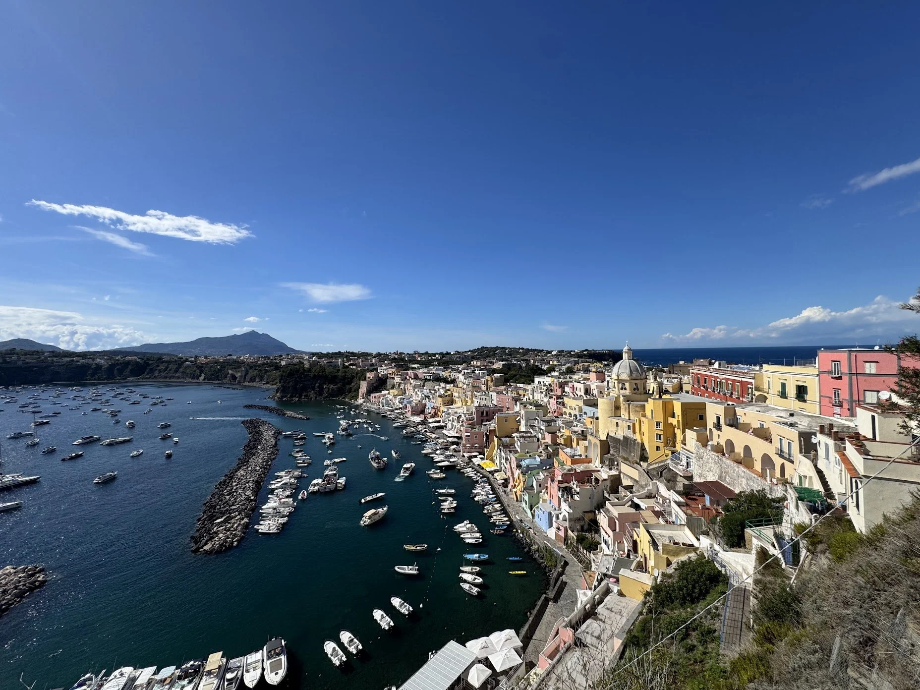 Colorful buildings along the coast with a marina filled with boats and a breakwater in a seaside town, under a partly cloudy blue sky.
