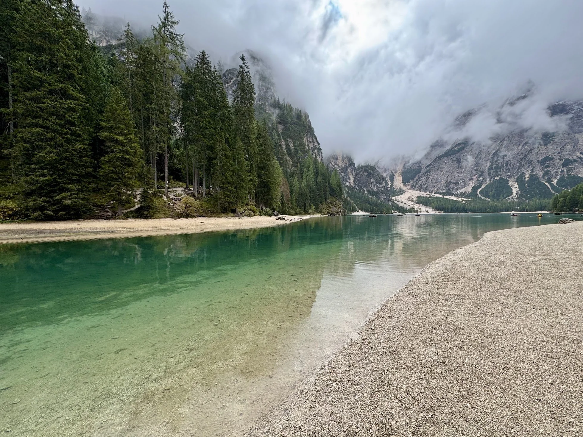 Scenic view of a calm green lake with mountain backdrop, dense green pine forest on the left, overcast sky with clouds and mist.