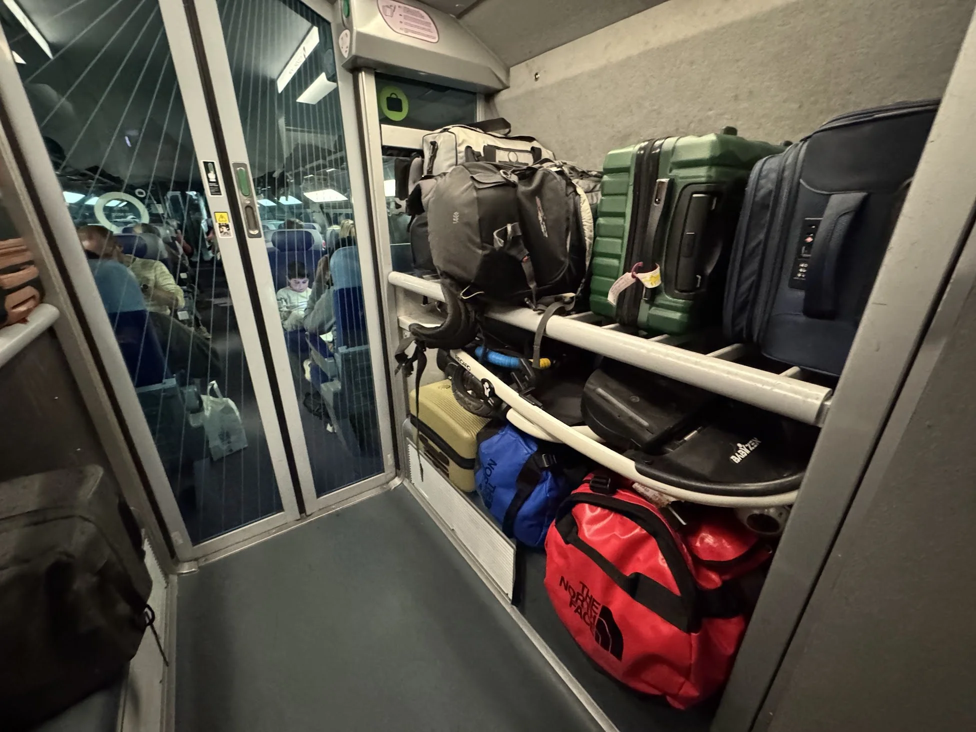 Luggage and backpacks stored on a luggage cart at an airport gate, with passengers seated in the background.