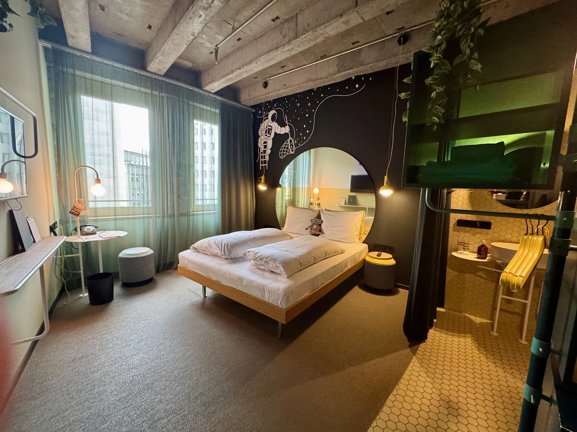 Interior of a modern hotel room with a bed, side table, desk, and a wall-mounted TV. Decor includes a black wall with an astronaut mural, green curtains over windows, and yellow accent lighting. Exposed concrete ceiling and some indoor plants are vis