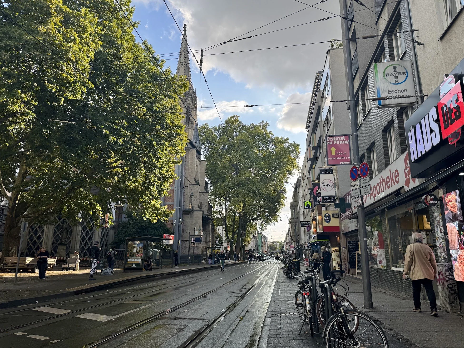 City street scene with tram tracks, people walking, bicycles parked, and storefronts with signs, with a church tower and trees in the background on a partly cloudy day.