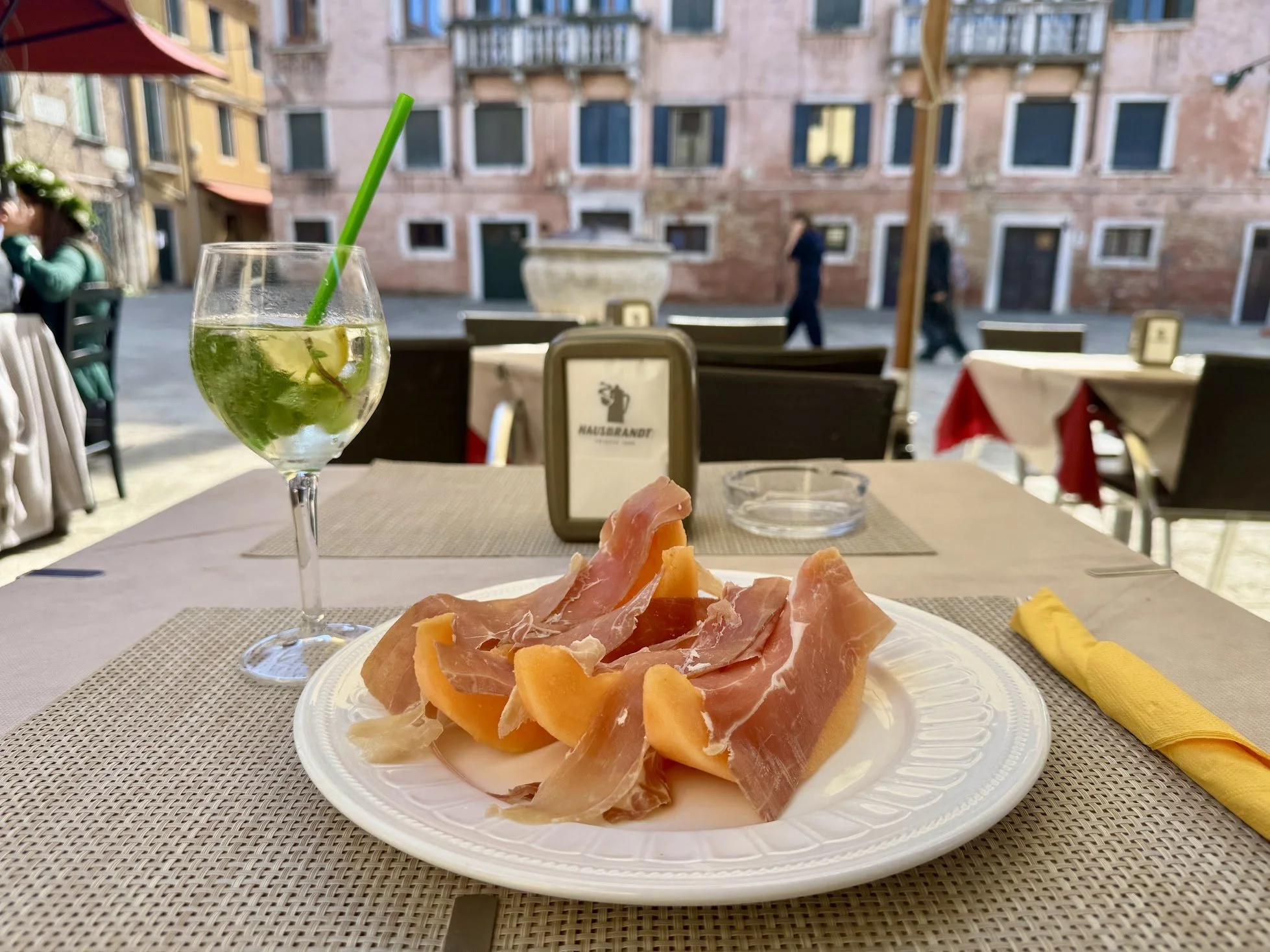 A plate with slices of prosciutto and cantaloupe on an outdoor restaurant table. In the background, a glass of green beverage with ice and a straw, and a view of buildings and people walking.