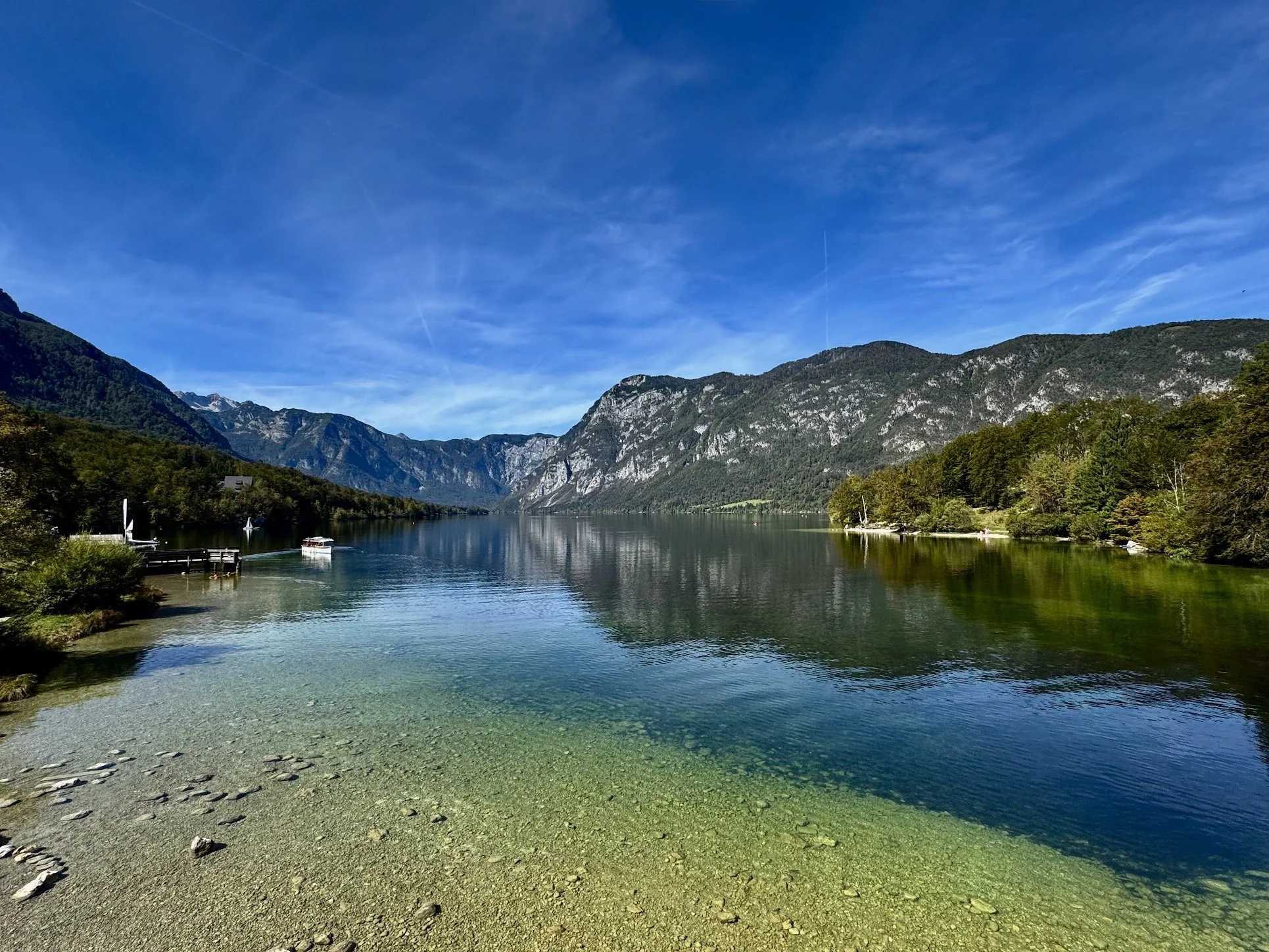 Scenic view of a calm river surrounded by lush green trees and towering mountains under a bright blue sky with a few wispy clouds.