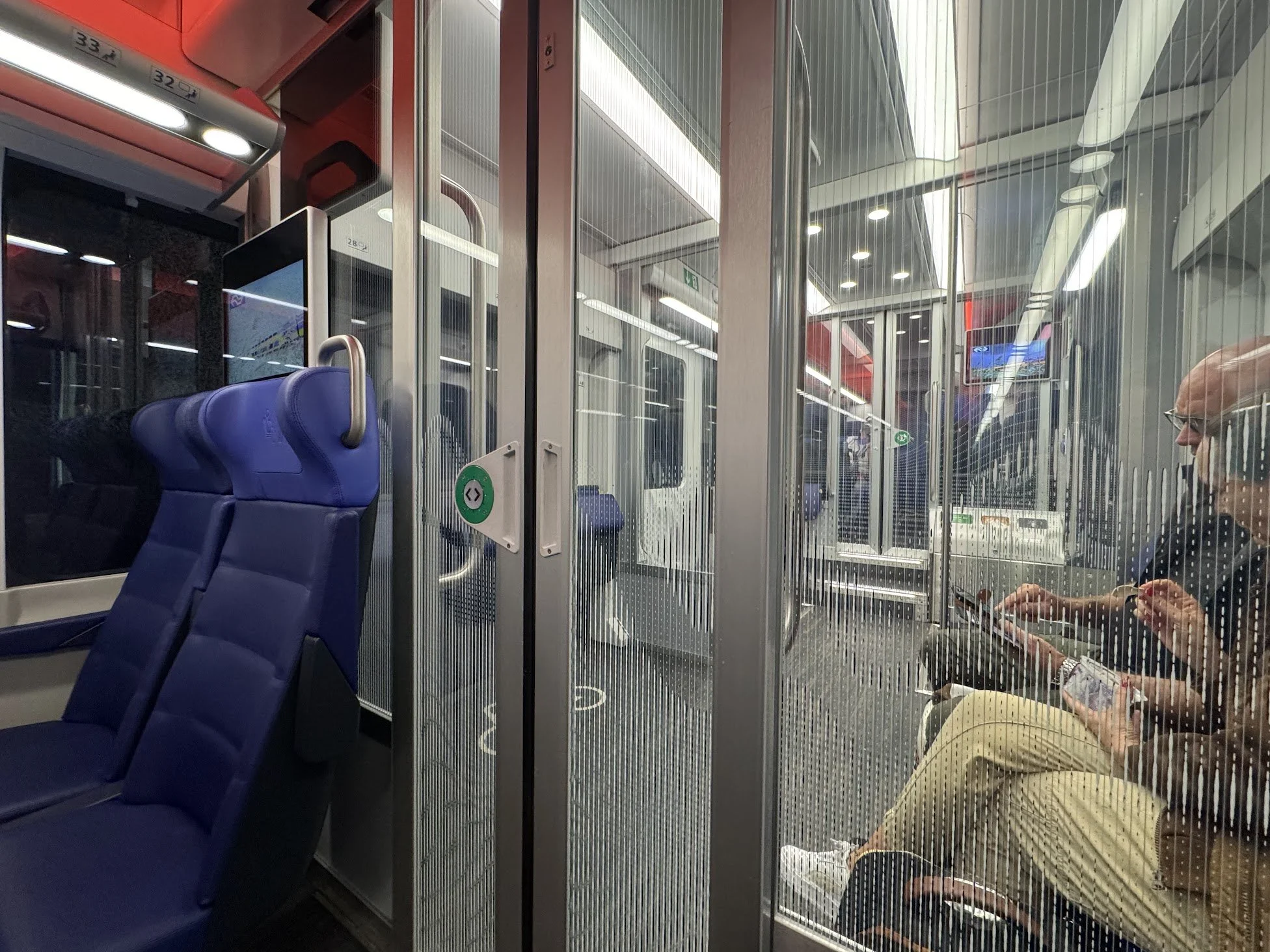 Interior of a bus or train with blue seats, glass dividers, and a woman sitting and looking at her phone.