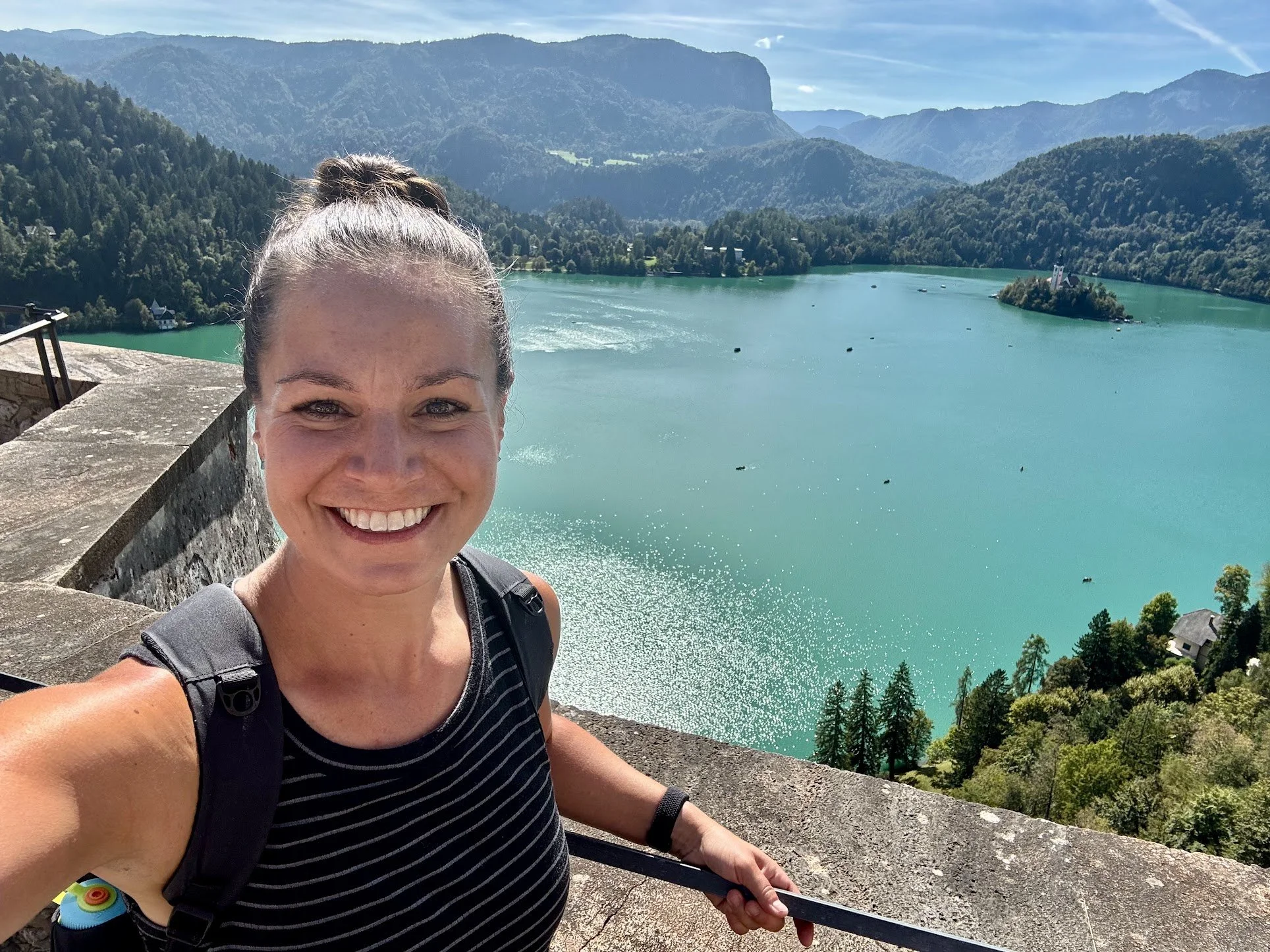 A woman with a ponytail smiling at the camera, standing on a stone ledge with a scenic lake and mountains in the background, wearing a black and white striped shirt and carrying a backpack.